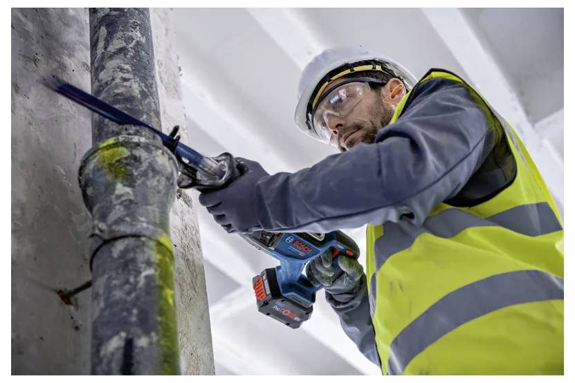 Ein Bauarbeiter in einer gelben Warnweste und Schutzhelm benutzt ein Elektrowerkzeug, um ein Rohr zu durchtrennen. Die Umgebung deutet auf eine industrielle Umgebung hin.