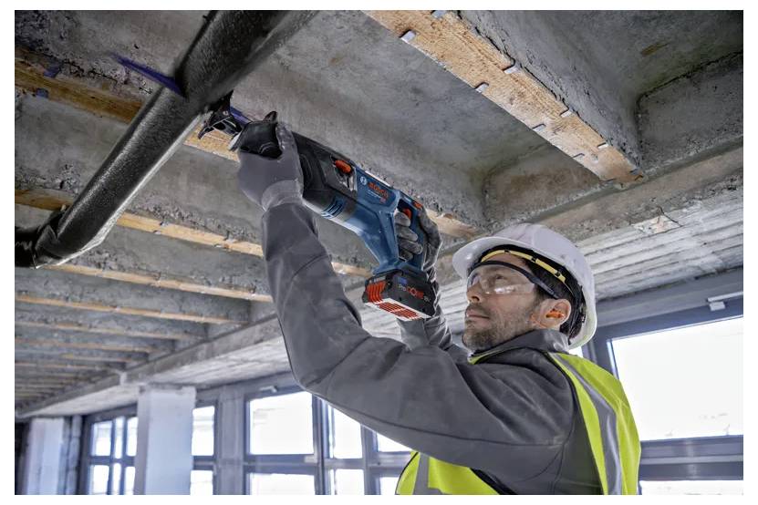Ein Bauarbeiter mit Schutzhelm und Warnweste verwendet ein Elektrowerkzeug, um eine Betondecke zu bohren, wobei sichtbare Rohre und Träger im Hintergrund sind.
