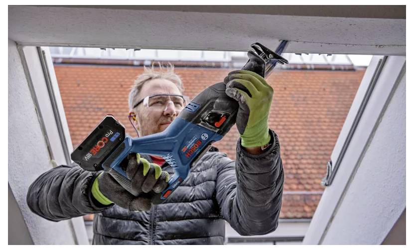 Eine Person mit Handschuhen und Schutzbrille verwendet eine Kreissäge, um eine Struktur zu durchtrennen, während im Hintergrund ein Dach zu sehen ist.