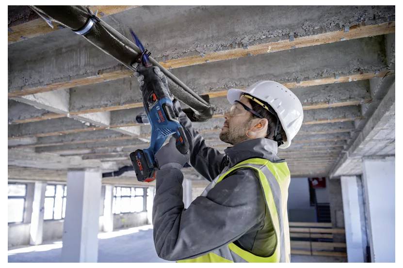 Ein Bauarbeiter mit Schutzhelm und Warnweste benutzt ein Elektrowerkzeug, um eine Deckenrohrleitung in einem unfertig ausgebauten Gebäudeinneren zu durchtrennen.