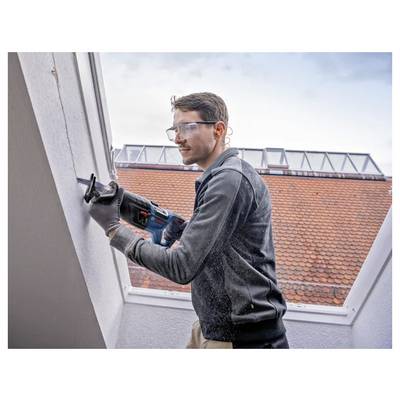 Ein Mann mit Sicherheitsbrille und Handschuhen verwendet ein Elektrowerkzeug, um eine Wand auf einem Dach zu schneiden, wobei orangefarbene Dachziegel im Hintergrund sichtbar sind.