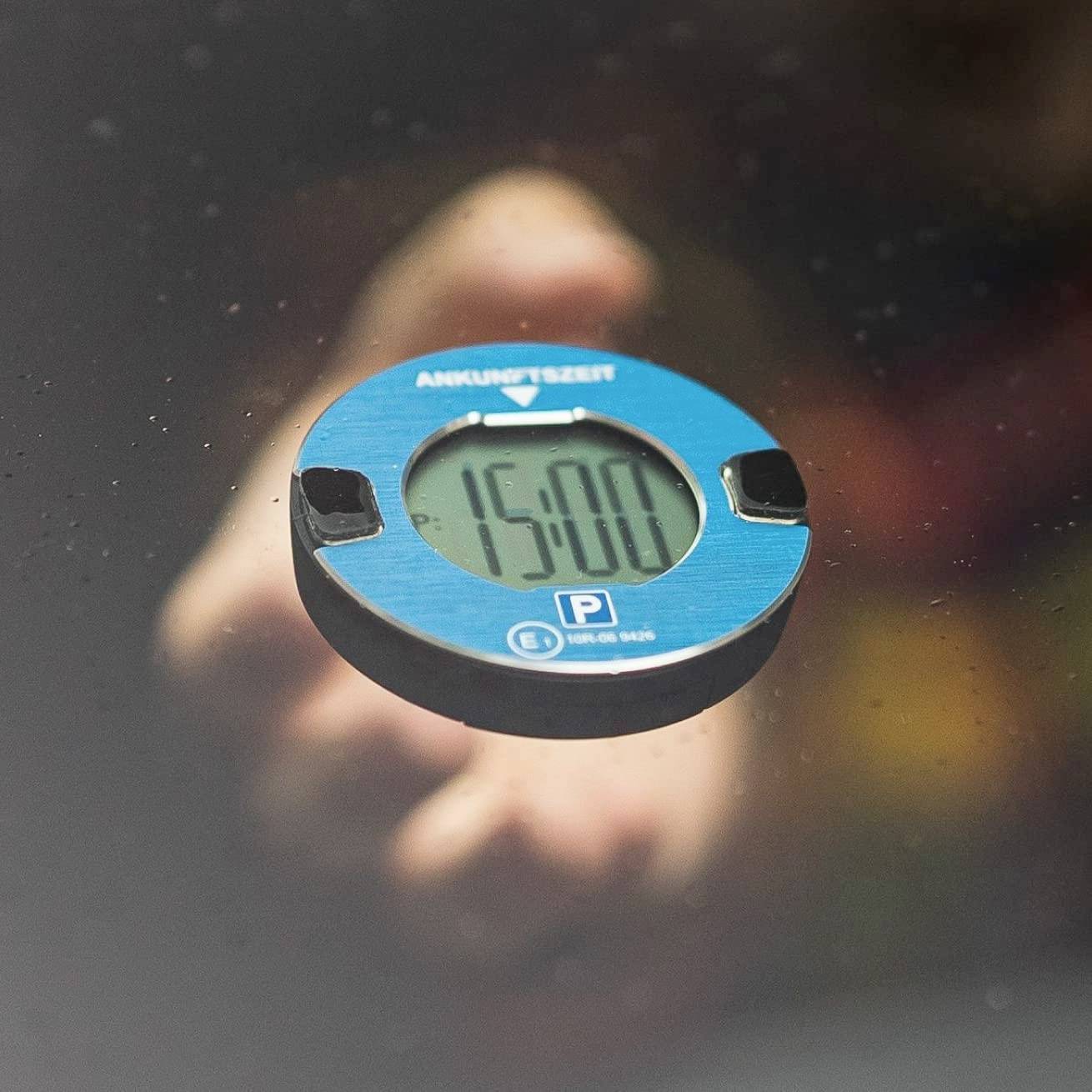 Eine blaue Parkscheibe zeigt die Ankunftszeit '15:00'. Sie wird von einer unscharfen Hand gehalten.