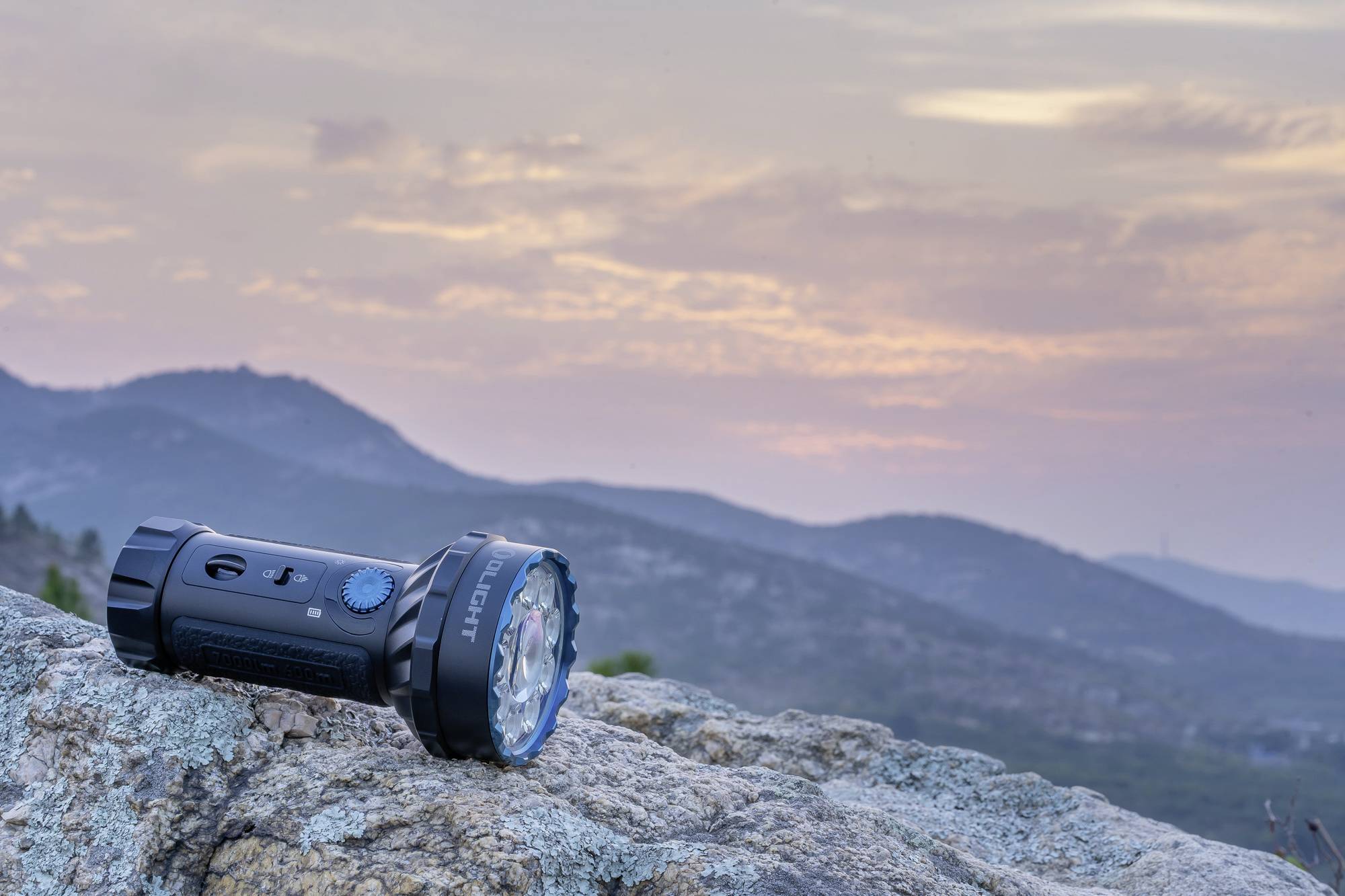 Eine schwarze Taschenlampe liegt auf einem Felsen im Vordergrund. Im Hintergrund sind Berge und ein farbenfroher Sonnenuntergang zu sehen.