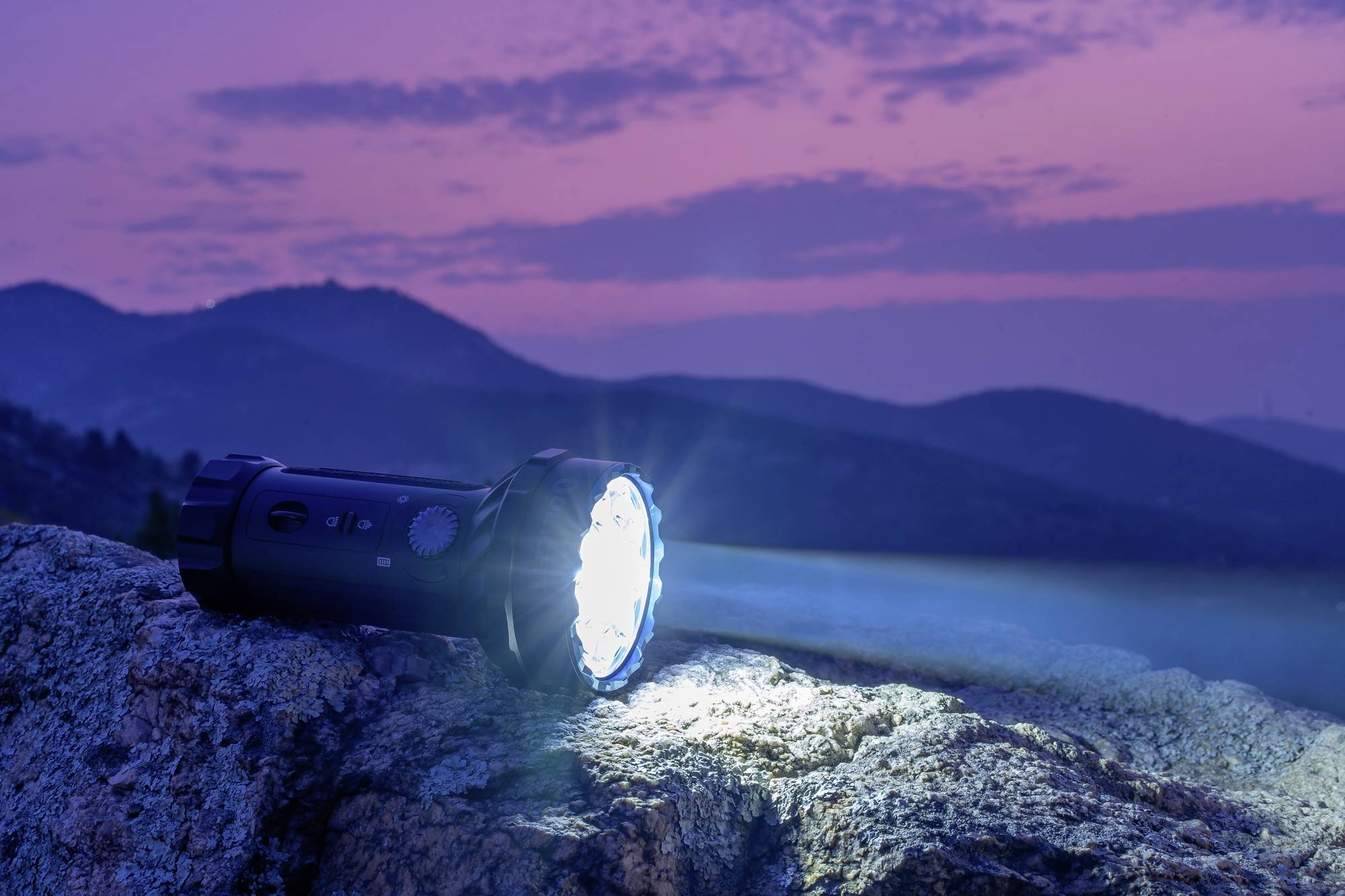 Taschenlampe leuchtet auf einem Felsen in der Dämmerung. Im Hintergrund sind hügelige Landschaft und violetter Himmel zu sehen.