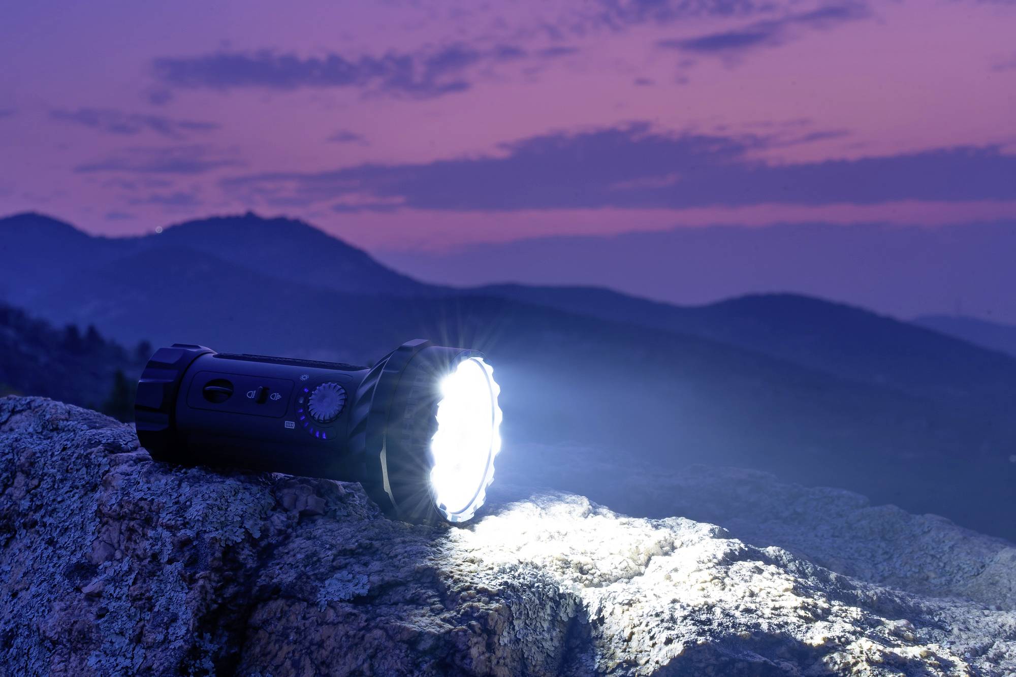 Taschenlampe leuchtet hell auf einem Felsen, im Hintergrund zeichnen sich Berge unter einem violetten Abendhimmel ab.
