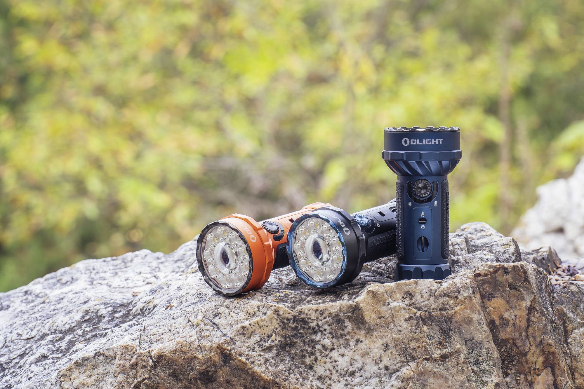 Zwei Taschenlampen, eine orange und eine schwarze, liegen auf einem Felsen im Freien. Hintergrund: unscharfe, grüne Vegetation.