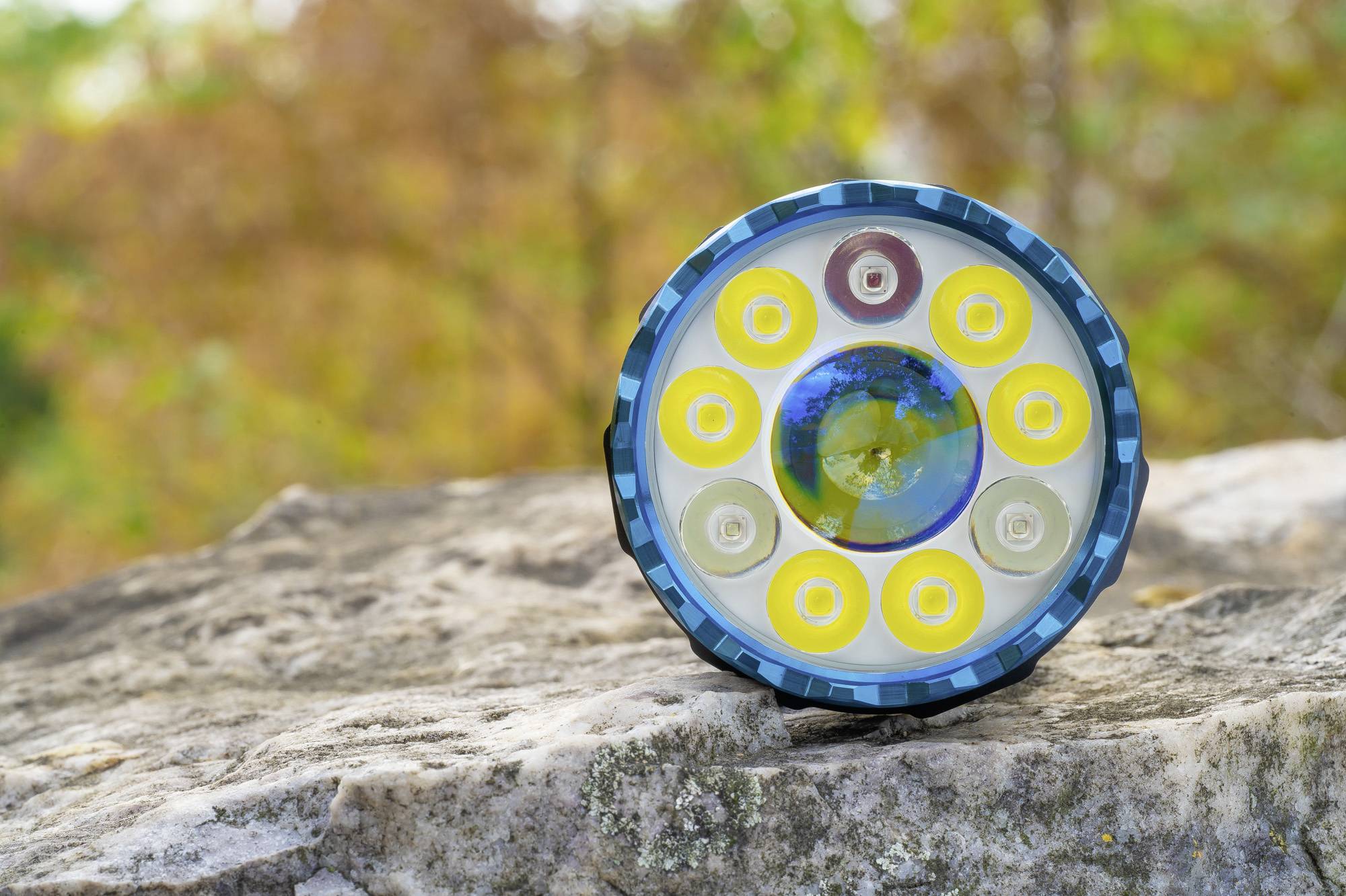 Blauer LED-Scheinwerfer auf einem Felsen im Freien, umgeben von grünen und herbstlichen Bäumen im Hintergrund. Fokus auf Gerätefront.