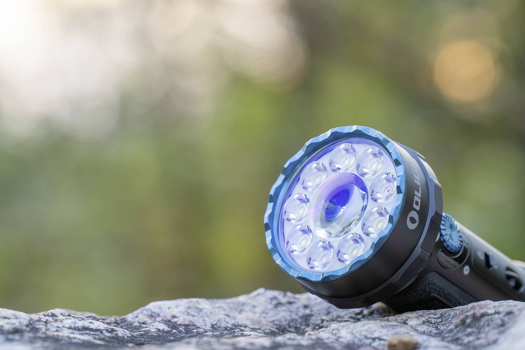 Eine Taschenlampe liegt auf einem Felsen, leuchtend mit mehreren LED-Lichtern. Im Hintergrund sind verschwommene Bäume sichtbar.