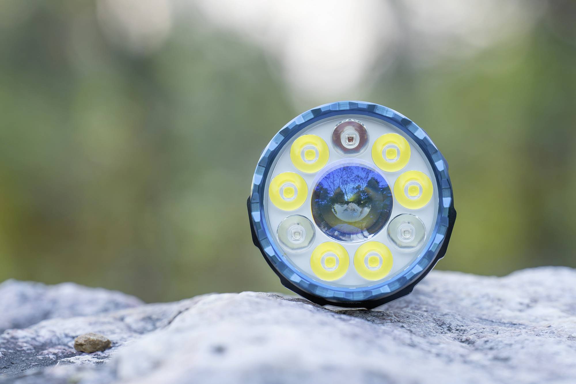 Nahaufnahme einer mehrfarbigen Taschenlampenlinse mit mehreren LED-Lichtern, auf einem Stein, verschwommener Waldhintergrund.