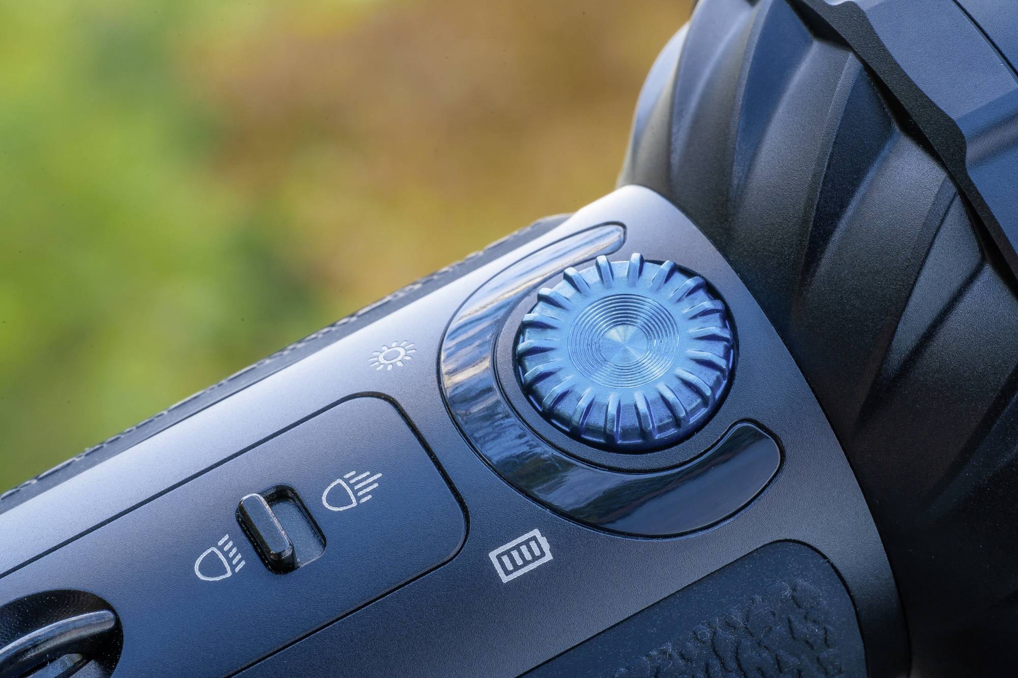 Nahaufnahme eines blauen Einstellrads an einer Taschenlampe, umgeben von weiteren Bedienelementen auf einem Hintergrund mit unscharfem Grün.
