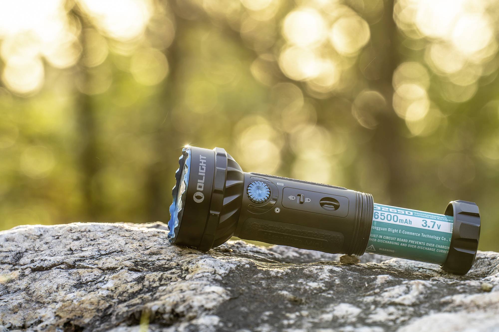 Eine schwarze Taschenlampe mit blauer Linse liegt auf einem Felsen, Hintergrund ist unscharf, goldenes Sonnenlicht leuchtet durch Bäume.