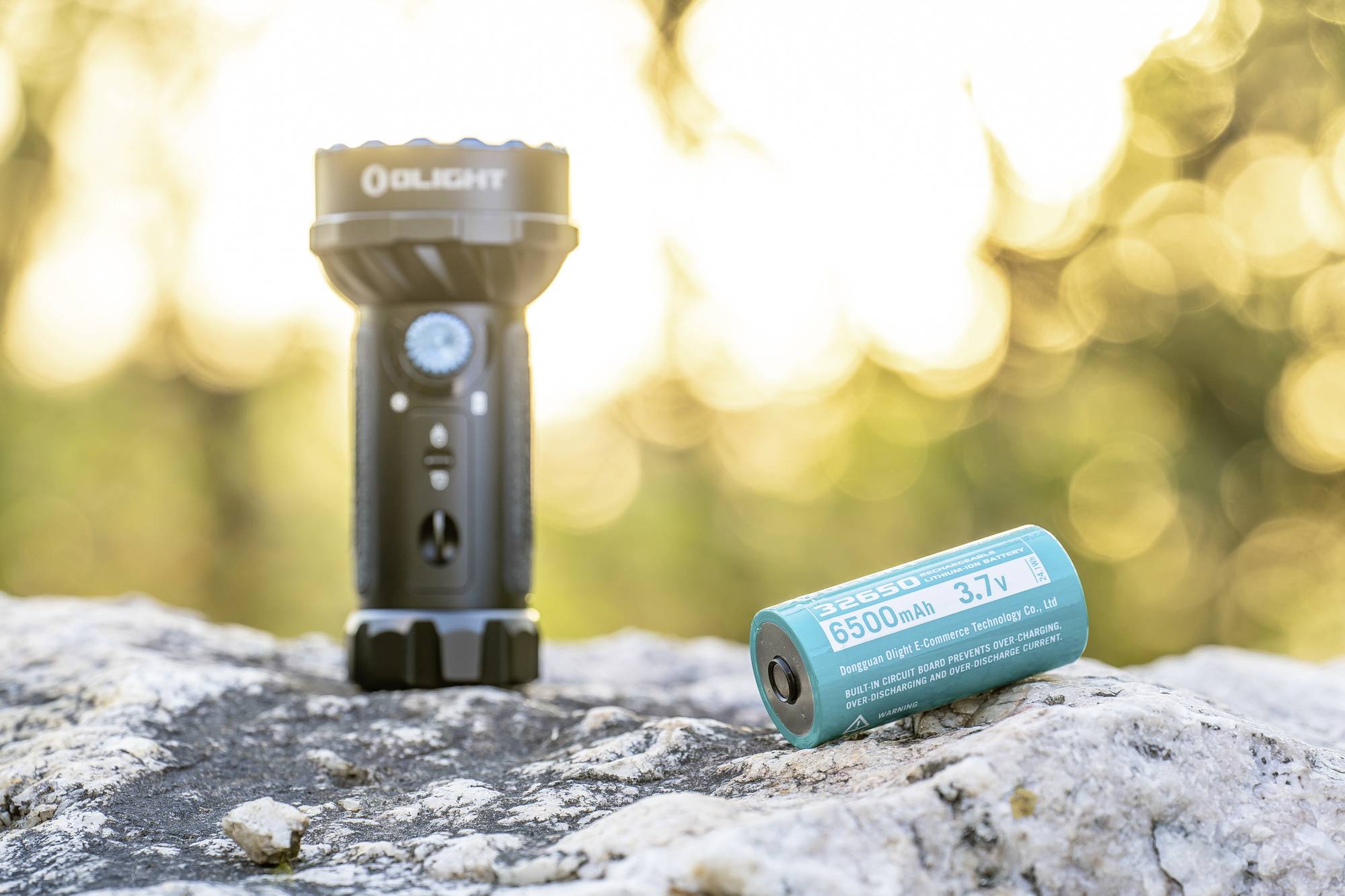 Taschenlampe und Batterie liegen auf einem Felsen im Freien. Hintergrund mit unscharfem, sonnendurchflutetem Wald.