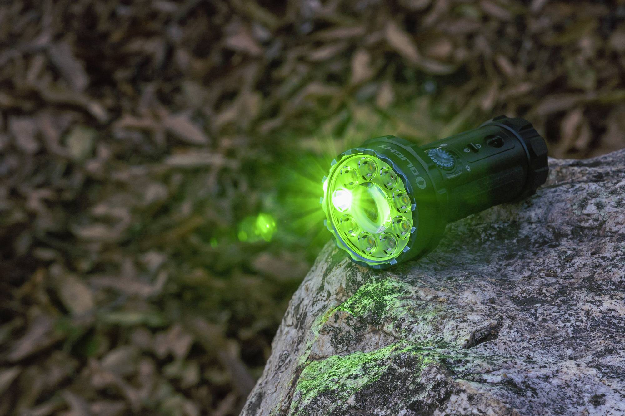 Eine Taschenlampe liegt auf einem Felsen und leuchtet mit grünem Licht. Im Hintergrund sind unscharf Blätter zu sehen.