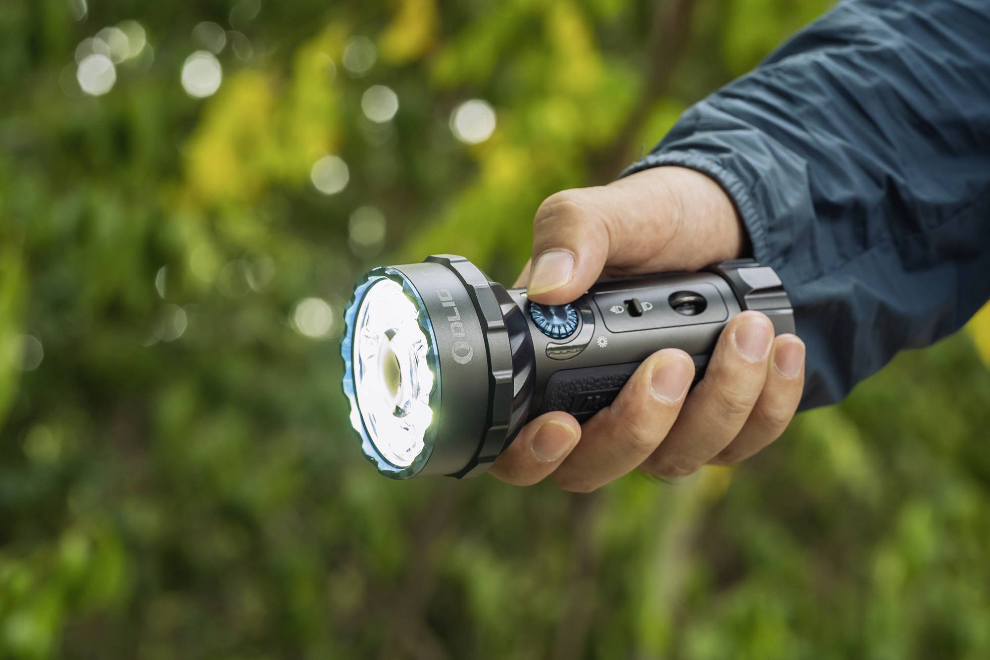Eine Hand hält eine leuchtende Taschenlampe, im Hintergrund grüne Bäume. Sie zeigt Nutzung im Freien bei schlechten Lichtverhältnissen.