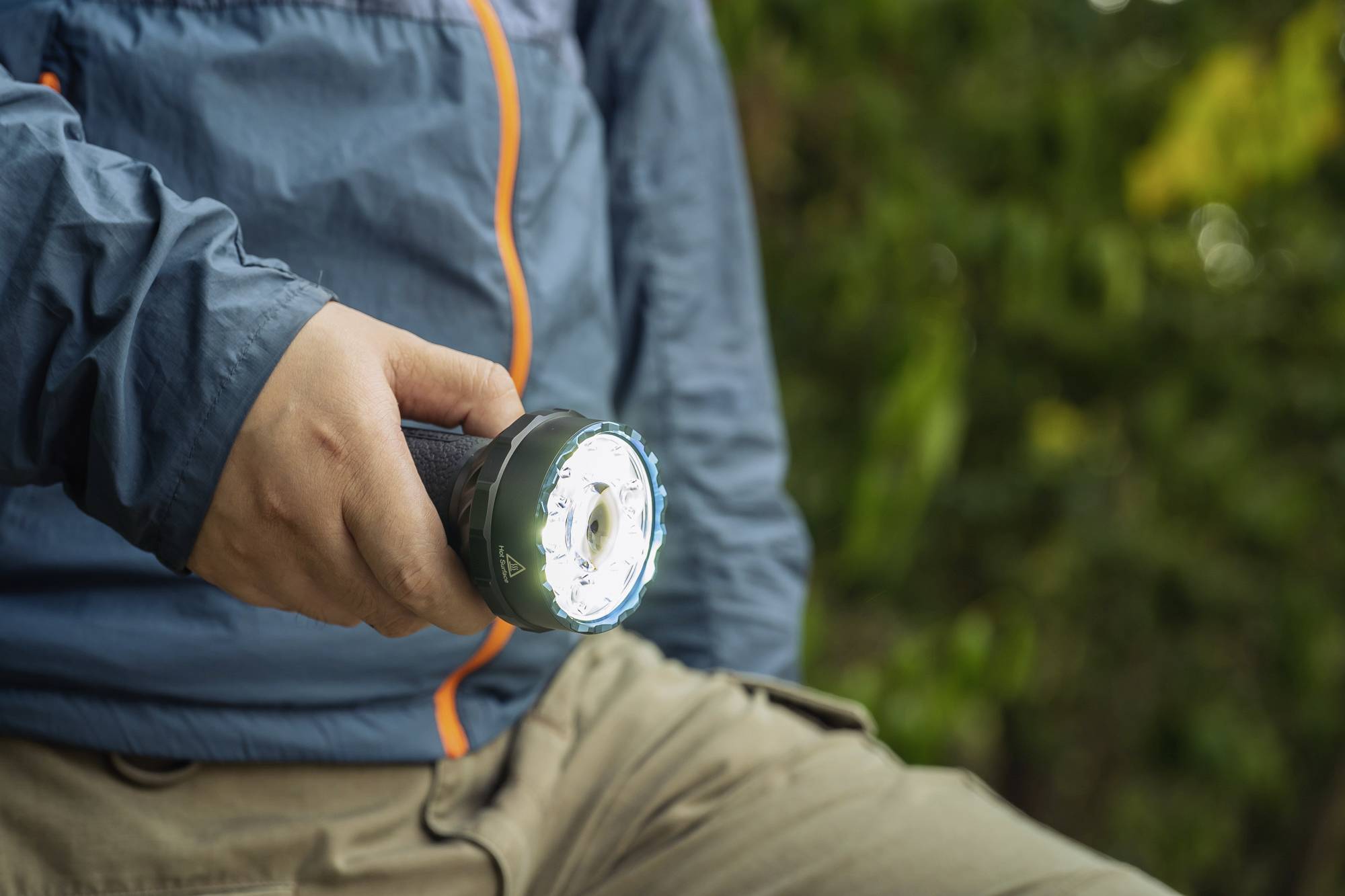 Person hält eine Taschenlampe in der Hand, zeigt sie nach vorne. Hintergrund unscharf grün. Person trägt Outdoor-Kleidung.