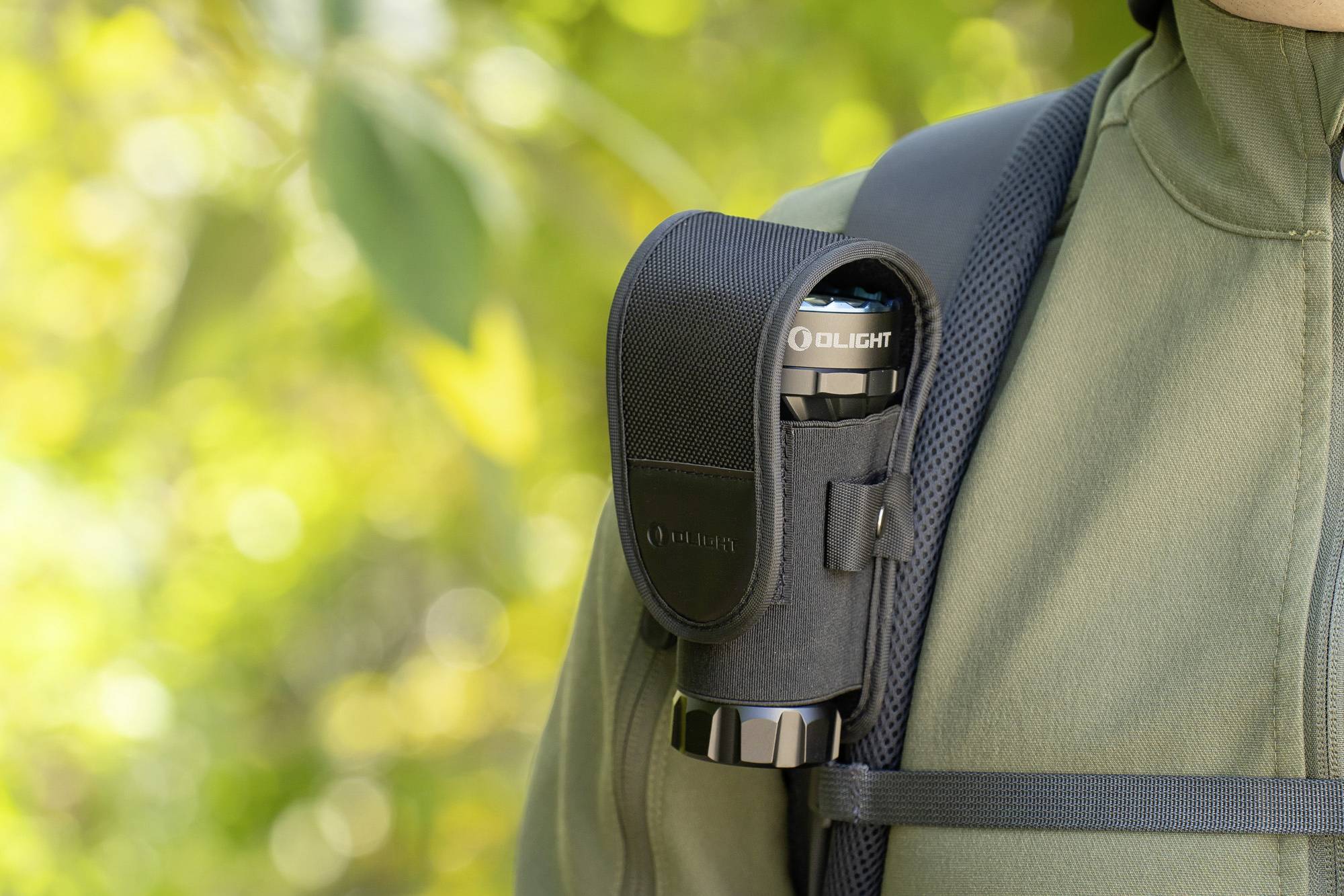 Person mit olivgrüner Jacke trägt einen Rucksack, an dem eine schwarze Taschenlampe an der Schulter befestigt ist. Im Hintergrund unscharfe Natur.