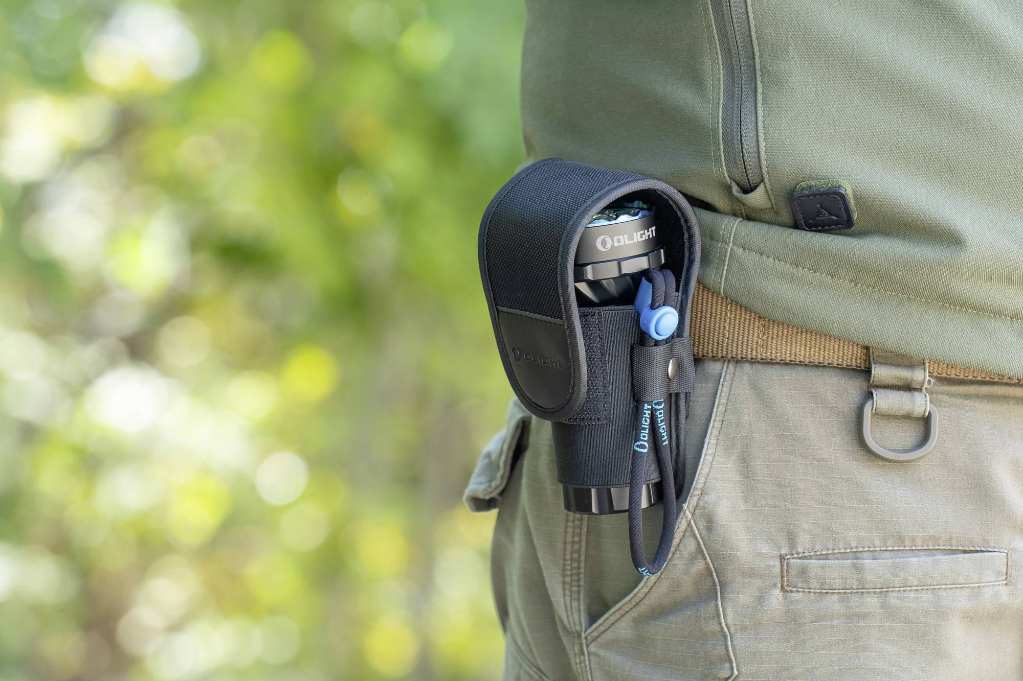 Eine Person trägt eine Taschenlampe in einer Halterung am Gürtel. Die Szene spielt in einer natürlichen, grünen Umgebung.