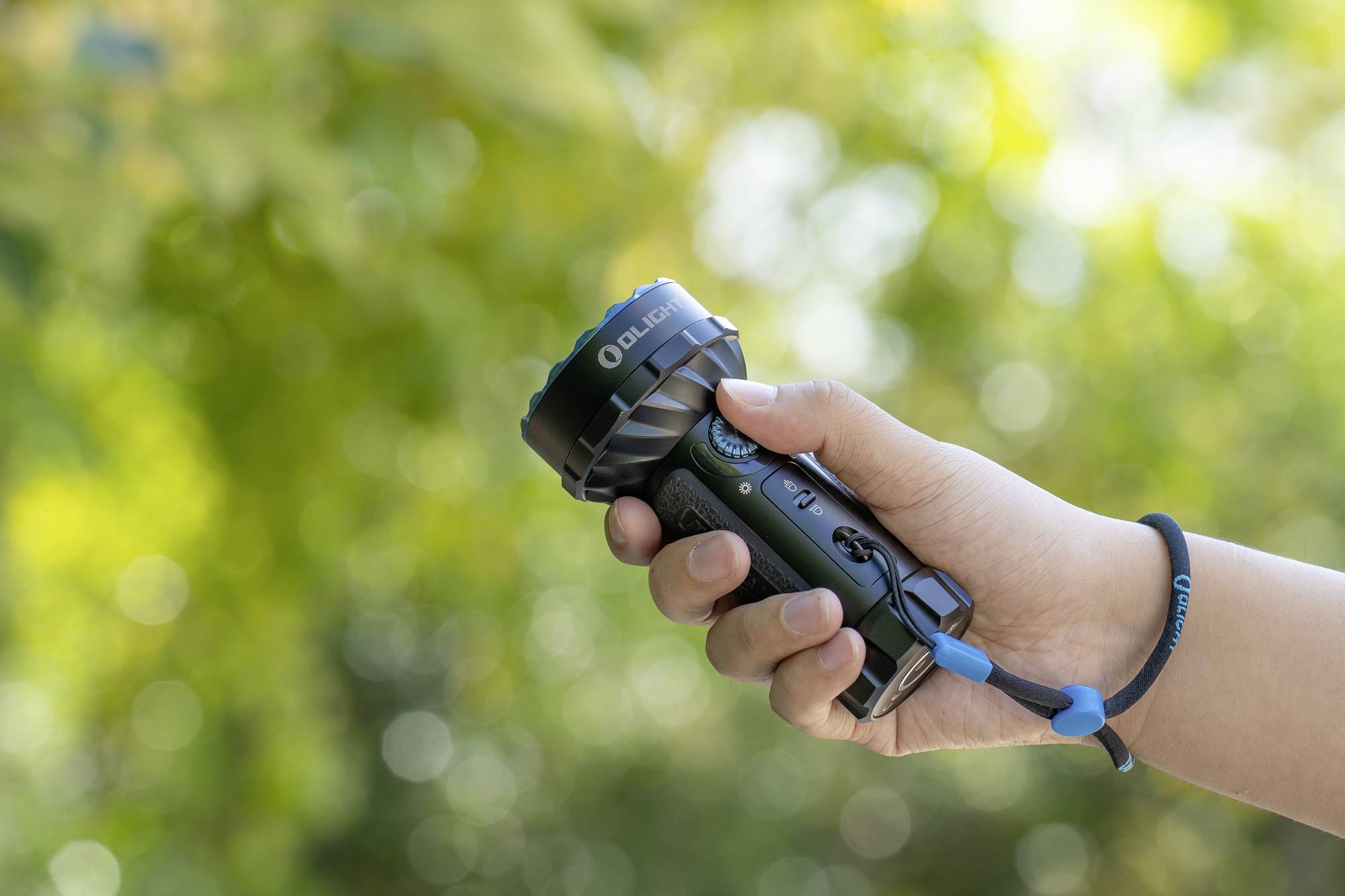 'Eine Hand hält eine Taschenlampe im Freien, umgeben von grünem, verschwommenem Laub im Hintergrund.'