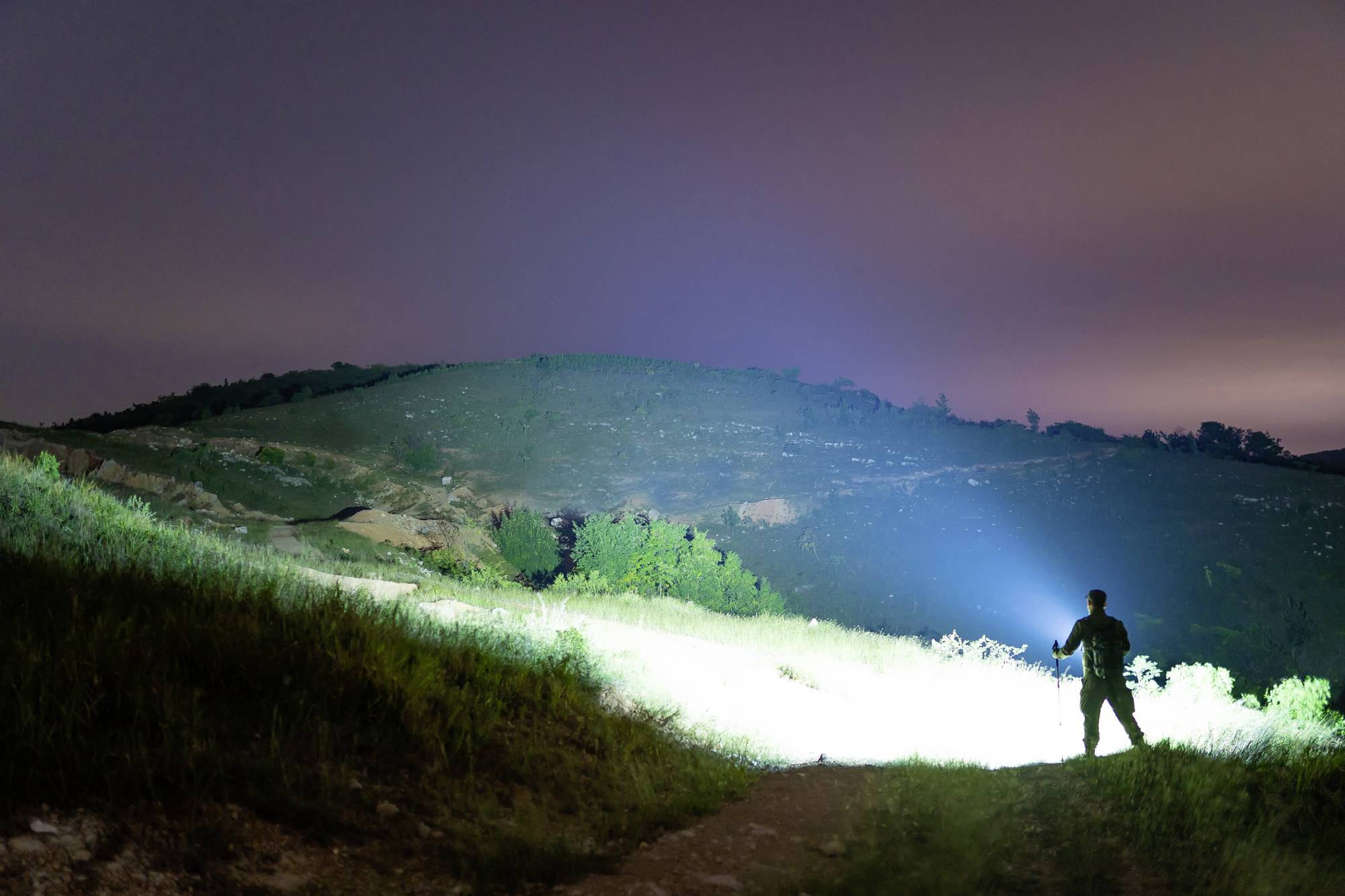 Eine Person steht in einem dunklen Feld und leuchtet mit einer Taschenlampe in die Ferne, beleuchtete Hügel im Hintergrund.