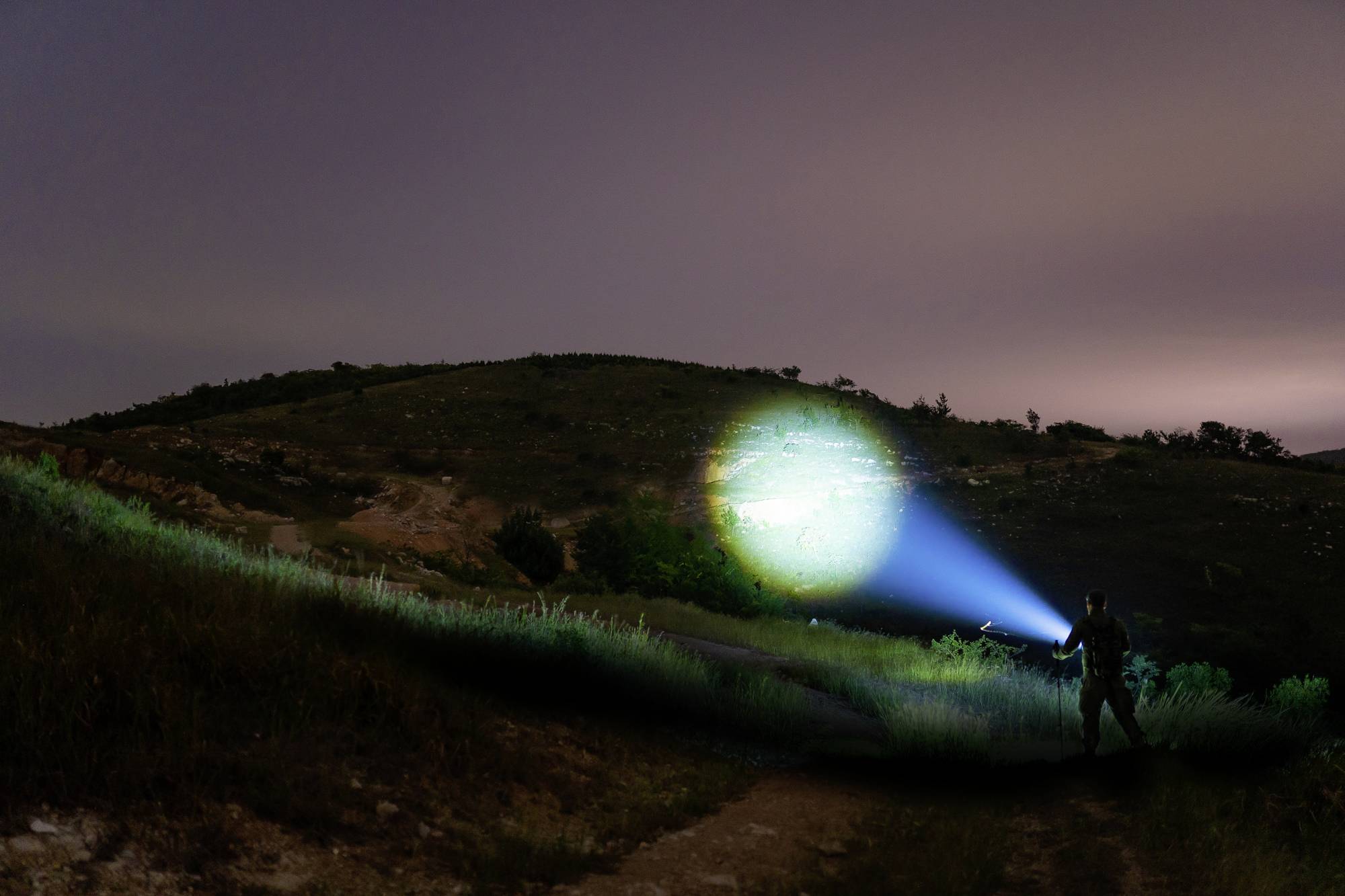Person mit Taschenlampe beleuchtet nächtliche Landschaft, Hügel im Hintergrund.