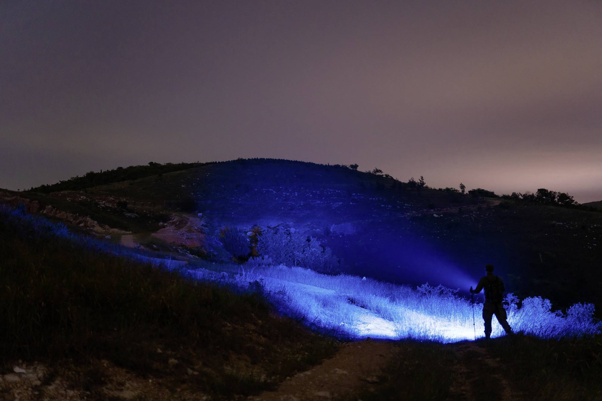 Ein Mensch steht auf einem Hügel in der Nacht und leuchtet mit einer hellen Taschenlampe, die blauen Schein auf den Boden wirft.