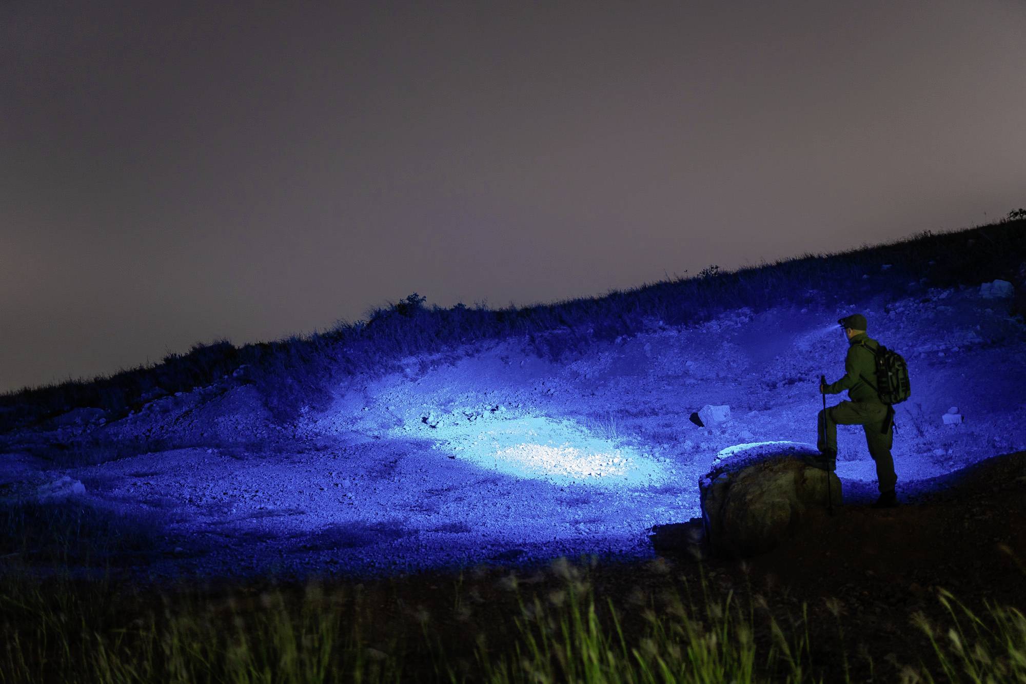 'Einsamer Wanderer steht bei Nacht auf einem Hügel, beleuchtet von mysteriösem blauem Licht im Hintergrund.'