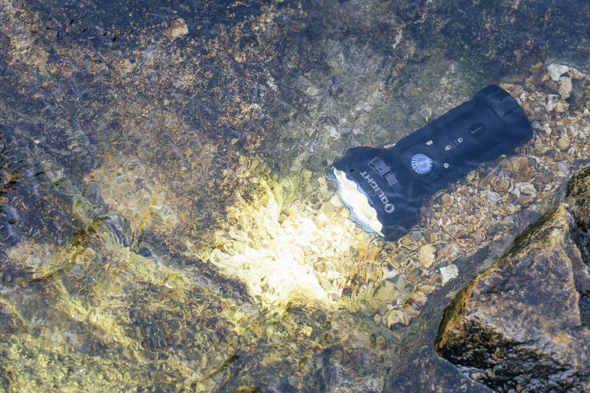 Eine wasserdichte Taschenlampe leuchtet unter Wasser auf einem flachen, steinigen Untergrund.