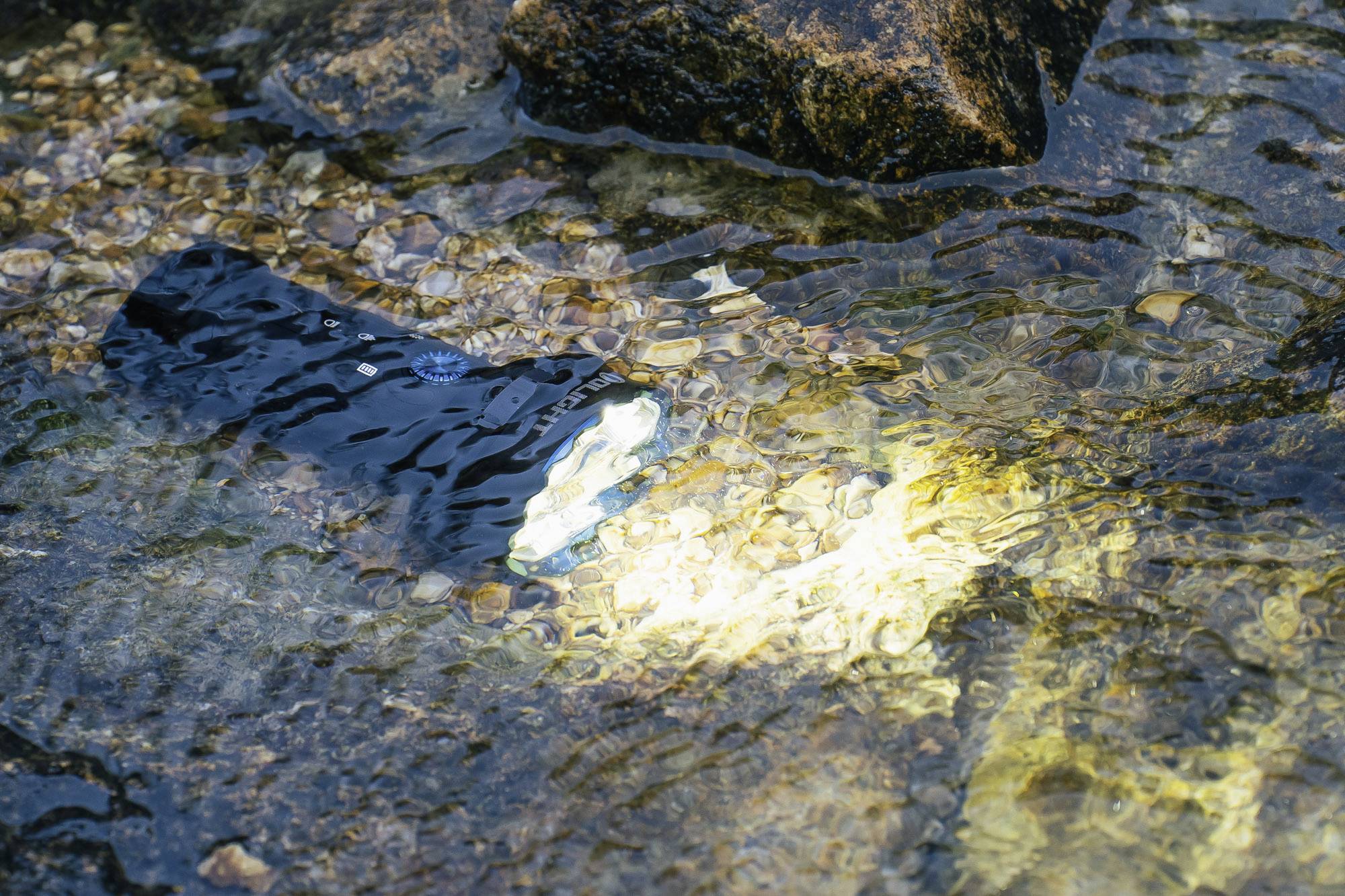 Taschenlampe strahlt unter Wasser auf, umgeben von Steinen. Licht und Schatten spielen auf der Wasseroberfläche.