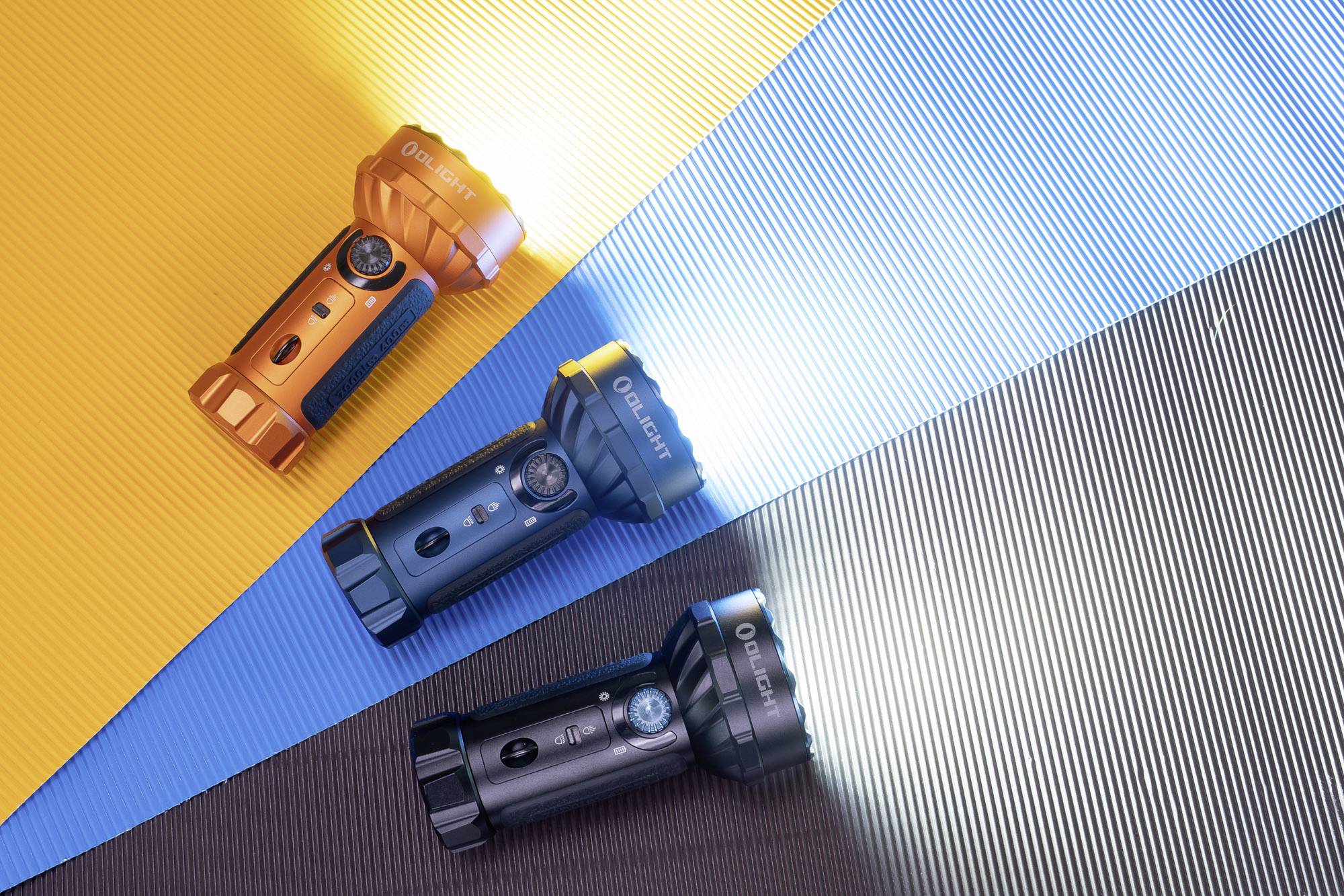 Drei Taschenlampen in Orange, Blau und Schwarz leuchten jeweils auf buntem, gestreiftem Hintergrund und bilden farbige Lichtkegel.