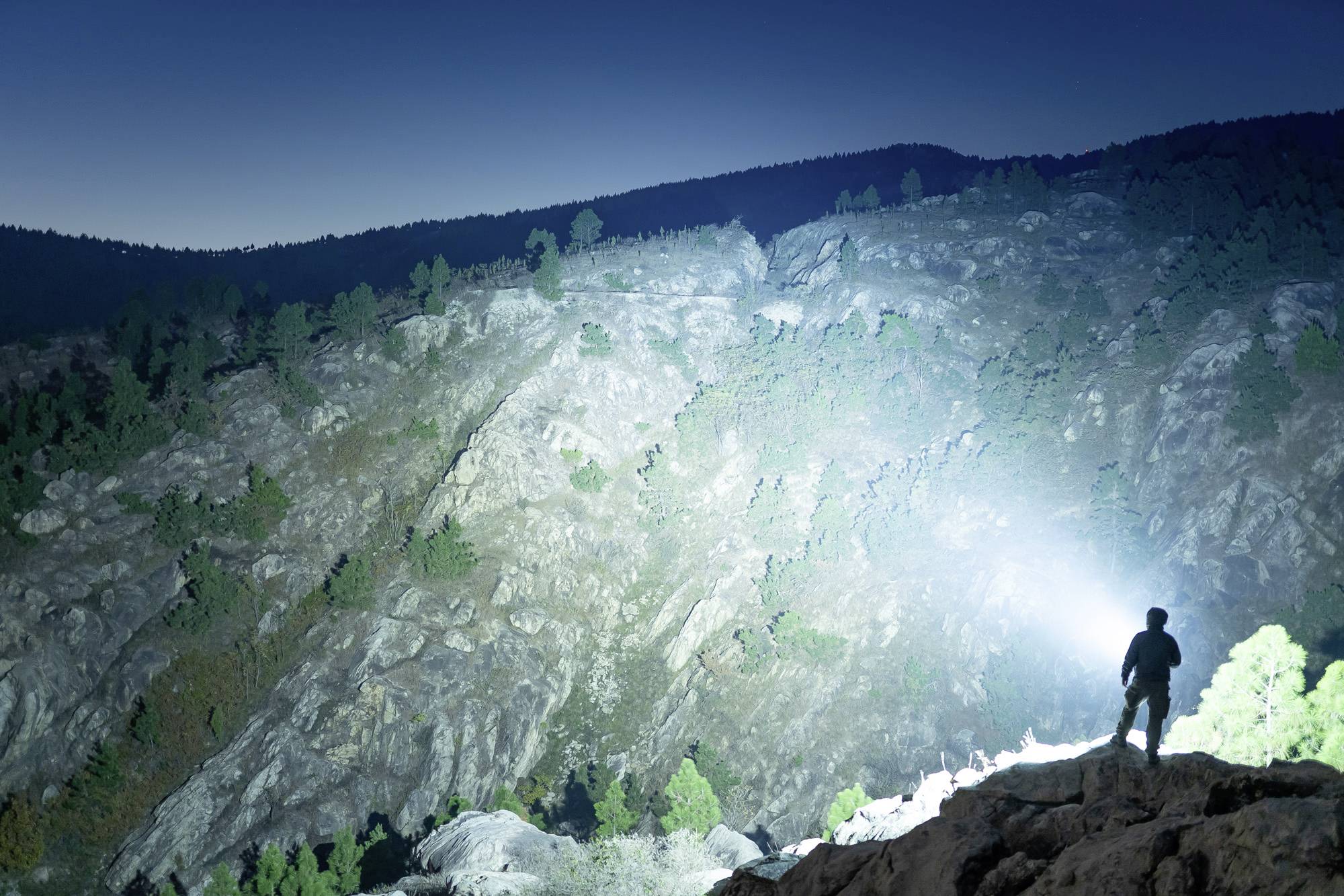Eine Person steht mit einer Taschenlampe auf einem Felsen in einer bergigen Landschaft, erleuchtet einen Teil des Hanges bei Dämmerung.