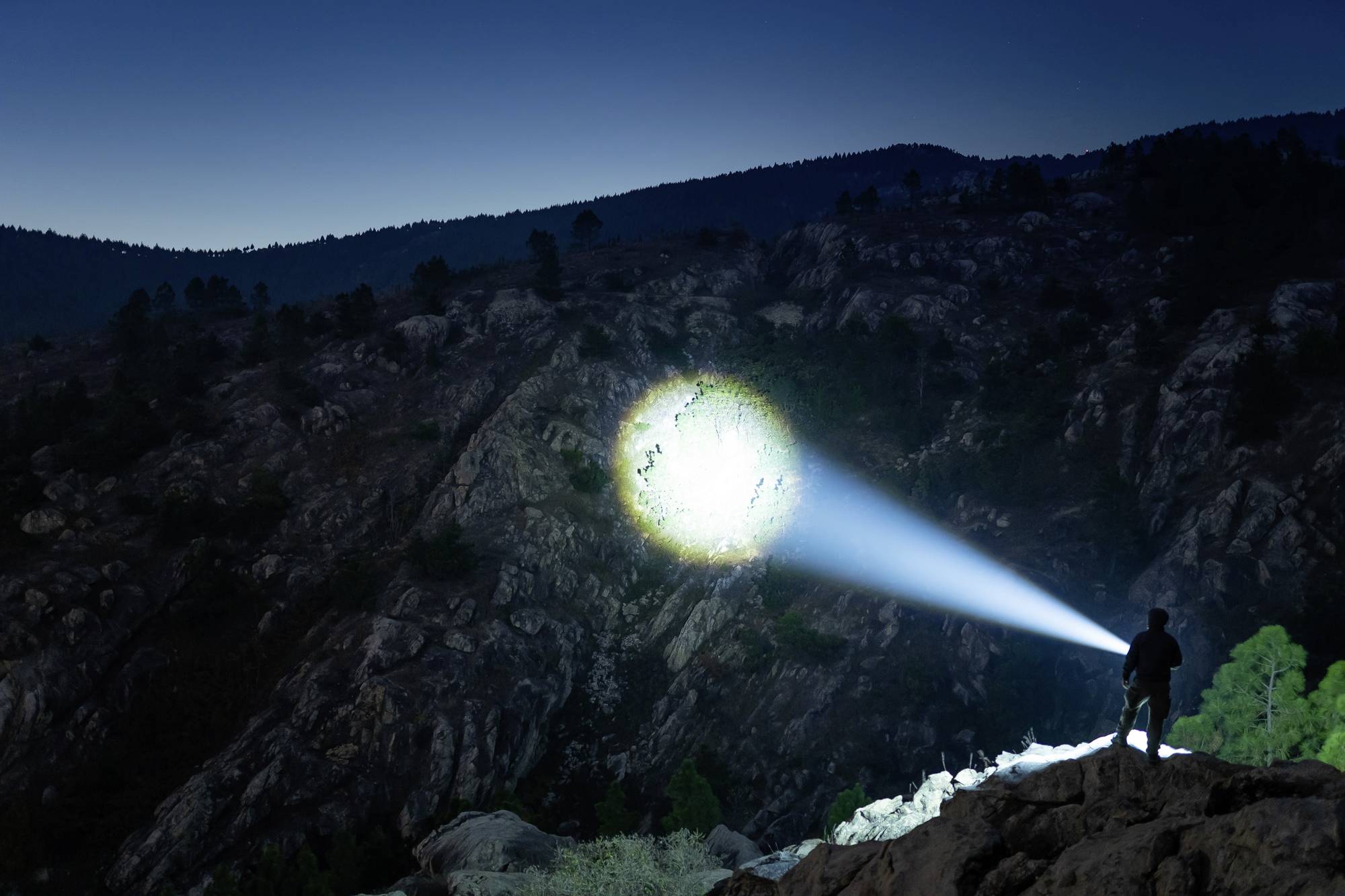 Ein Mensch steht auf einem Felsen bei Nacht und leuchtet mit einer starken Taschenlampe auf einen Berghang.