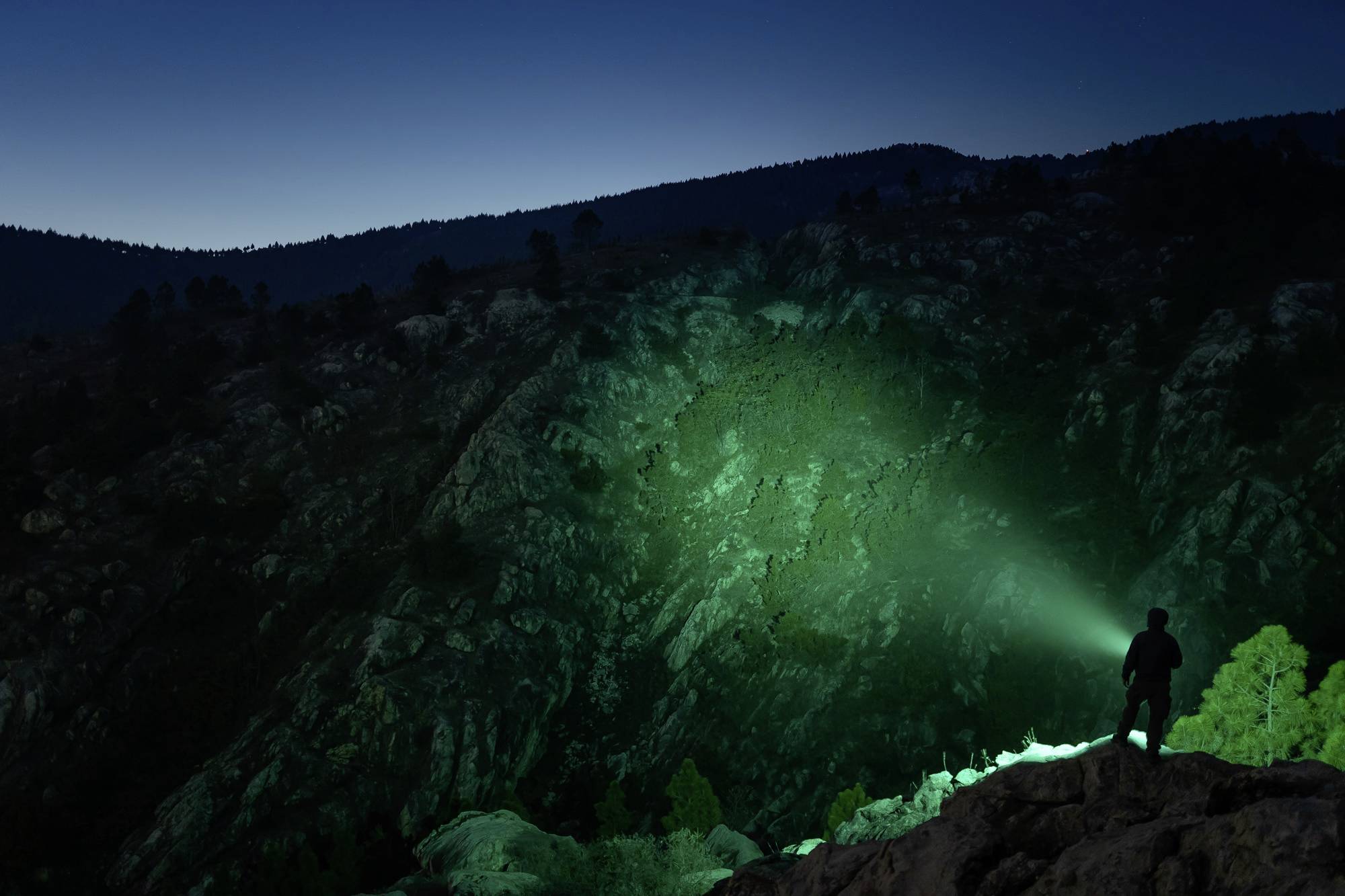 Eine Person steht nachts auf einem Felsen und leuchtet mit einer grünen Taschenlampe auf eine bewaldete Berglandschaft.