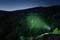 Eine Person steht nachts auf einem Felsen und leuchtet mit einer grünen Taschenlampe auf eine bewaldete Berglandschaft.