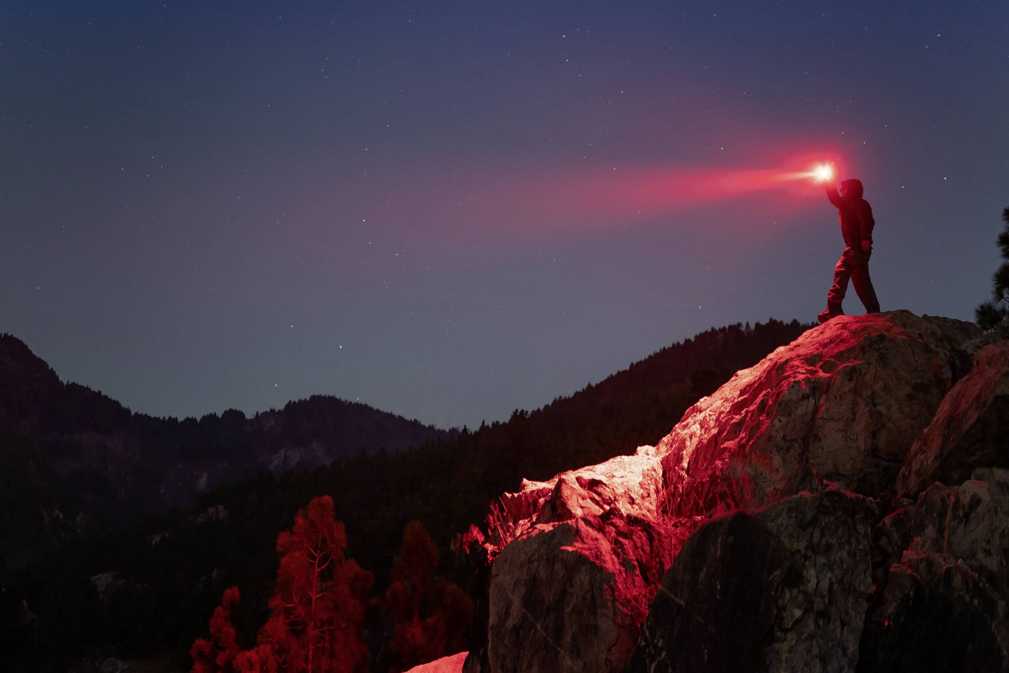 Eine Person steht auf einem Felsen und leuchtet mit einer roten Taschenlampe in die Nacht, umgeben von Bergen und einem klaren Himmel.