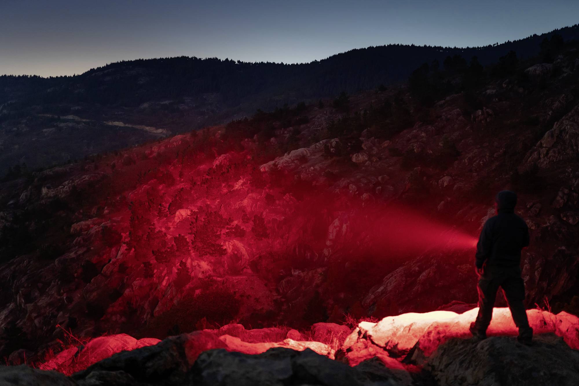 Eine Person steht nachts auf einem Felsen mit einem roten Scheinwerferlicht, das über die Landschaft schweift. Berge im Hintergrund.
