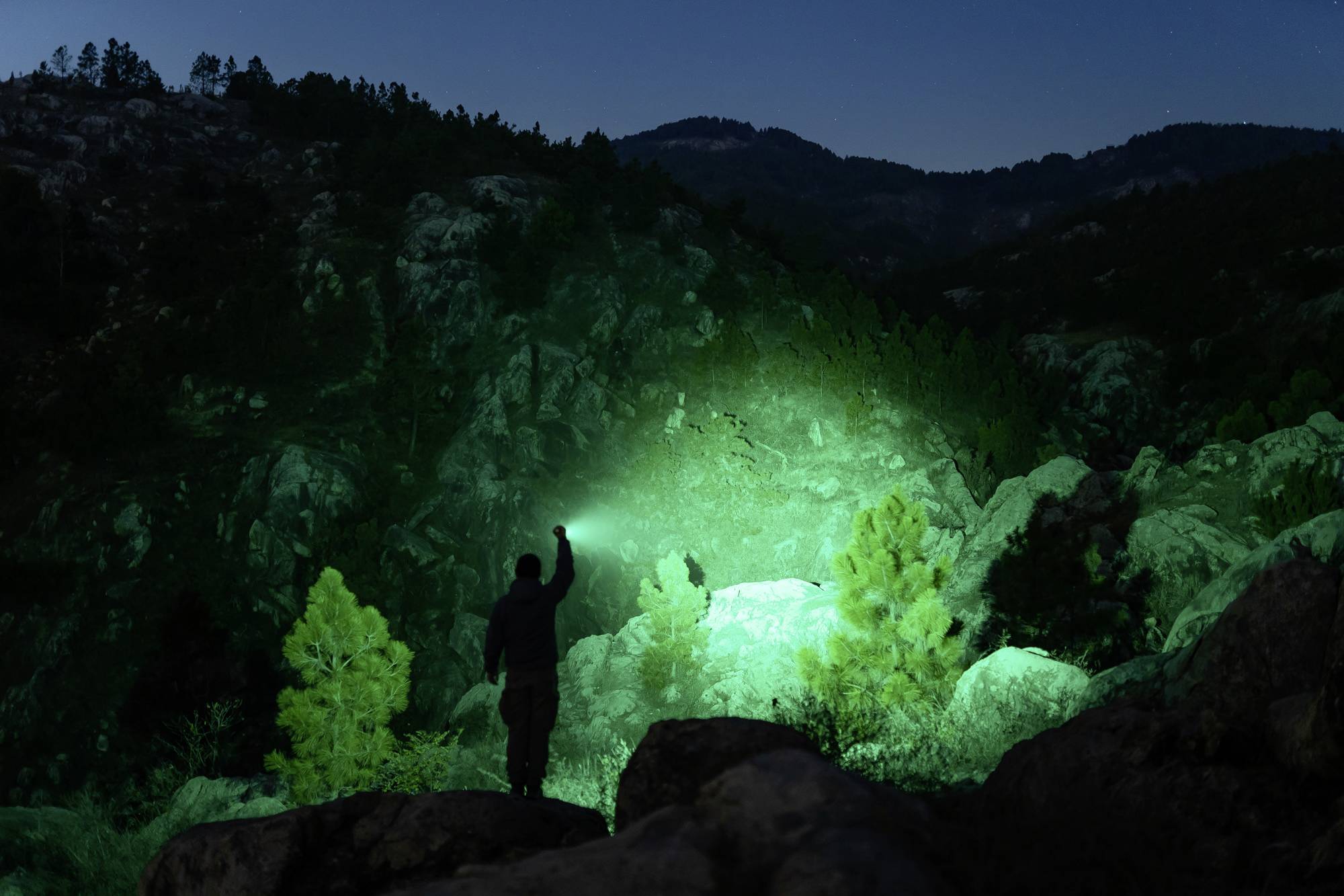 Eine Person steht nachts in einer felsigen Landschaft und leuchtet mit einer grünen Taschenlampe in die Dunkelheit.