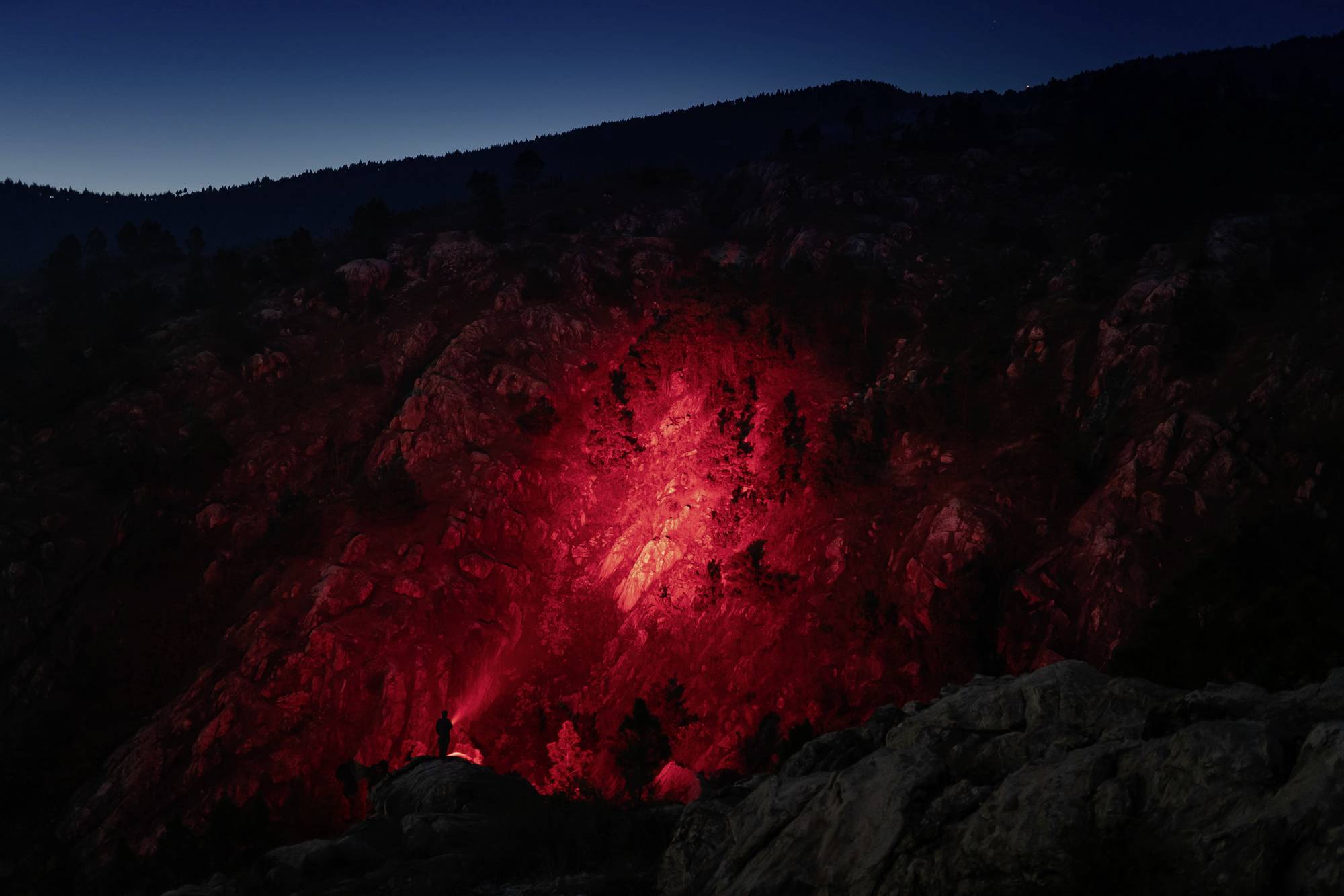 Eine dunkle Berglandschaft bei Nacht, beleuchtet von rotem Licht. Ein Mensch steht am unteren Bildrand mit einer Taschenlampe.