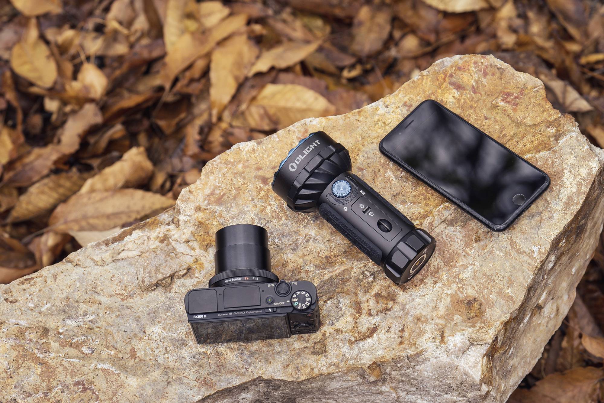 Kamera, Taschenlampe und Smartphone liegen auf einem großen Felsen im Freien, umgeben von herbstlichen Blättern.