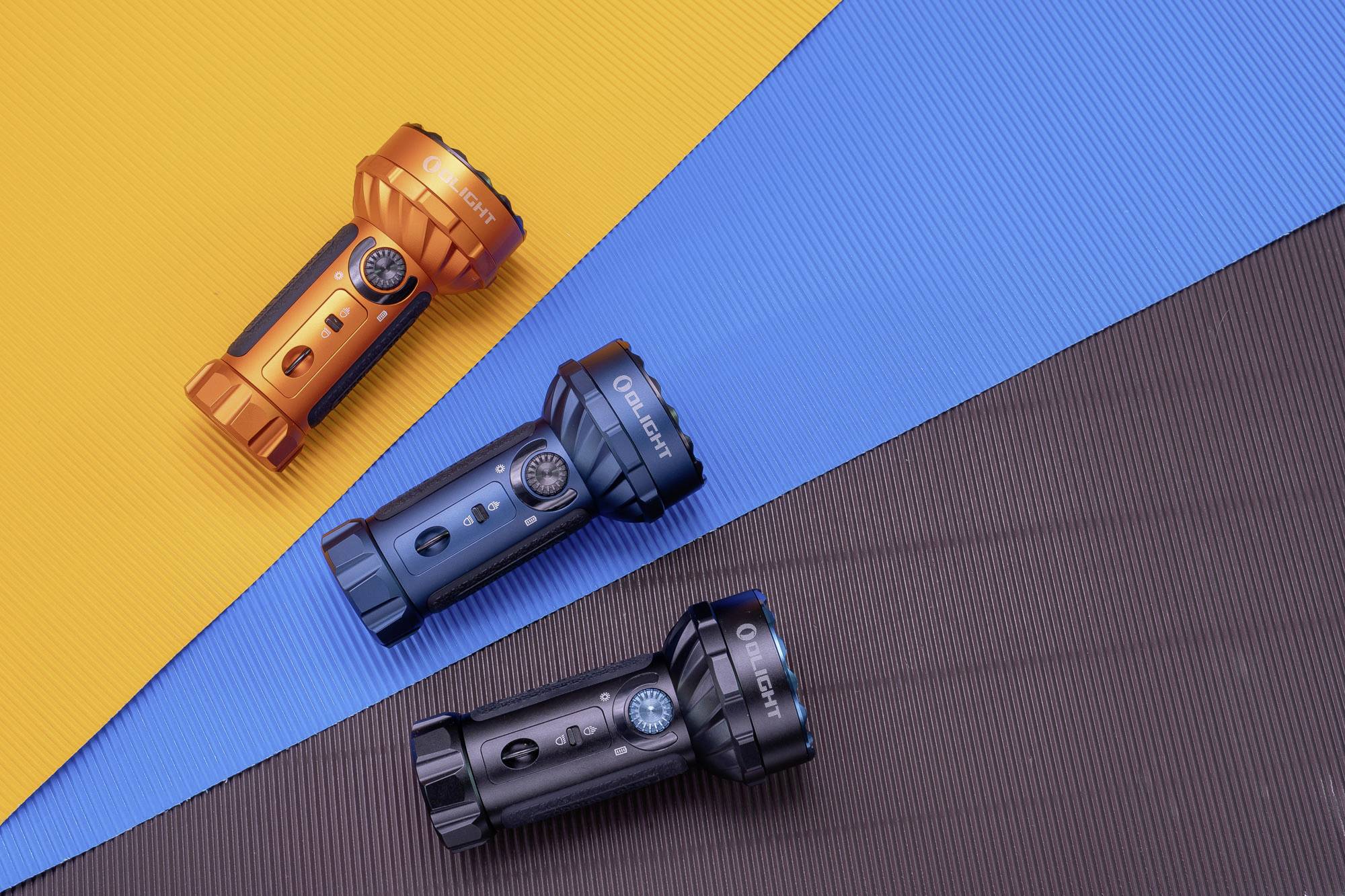 Drei Taschenlampen in Orange, Blau und Schwarz liegen diagonal auf farbigen Unterlagen in Gelb, Blau und Braun.