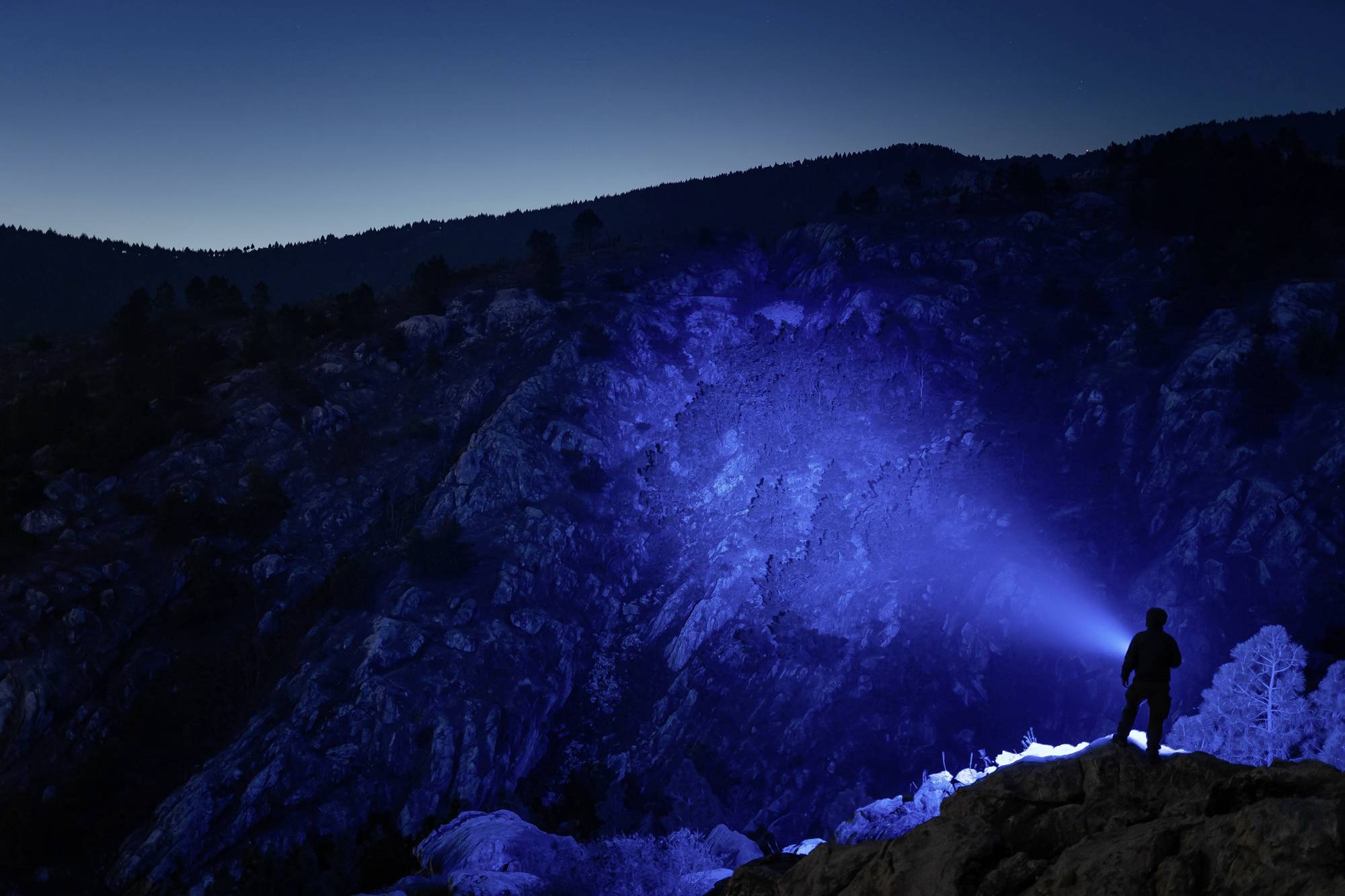 Eine Person steht auf einem Felsen bei Nacht und leuchtet mit einer Taschenlampe einen Berg an, wodurch ein blauer Lichtstrahl entsteht.