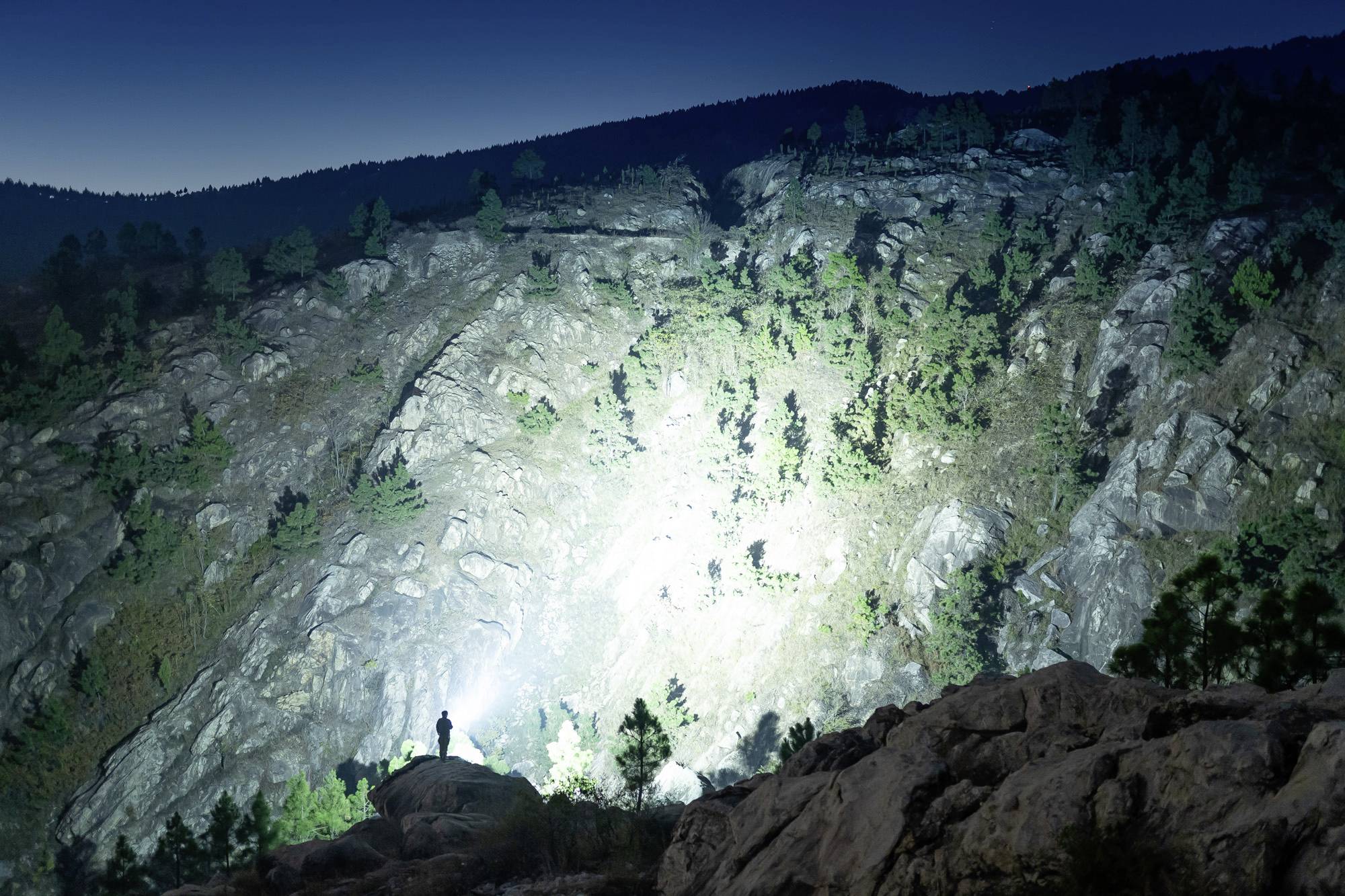 Eine Person steht nachts auf einem Felsen, beleuchtet von einem starken Licht, das auf eine bewaldete Bergwand im Hintergrund scheint.