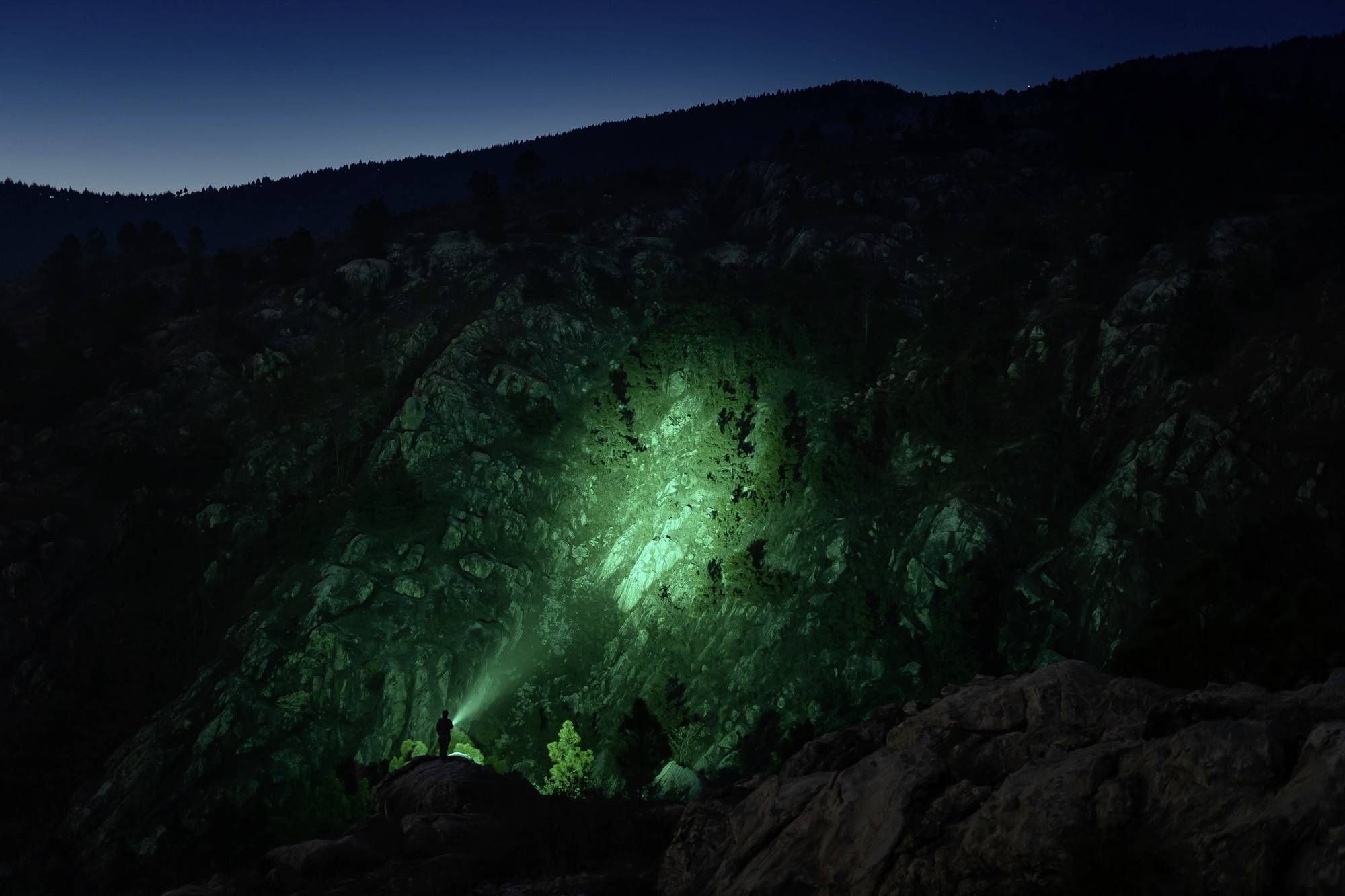 Berglandschaft bei Nacht mit grünem Licht, das Felsen beleuchtet. Eine Person steht im Vordergrund mit einer Taschenlampe.