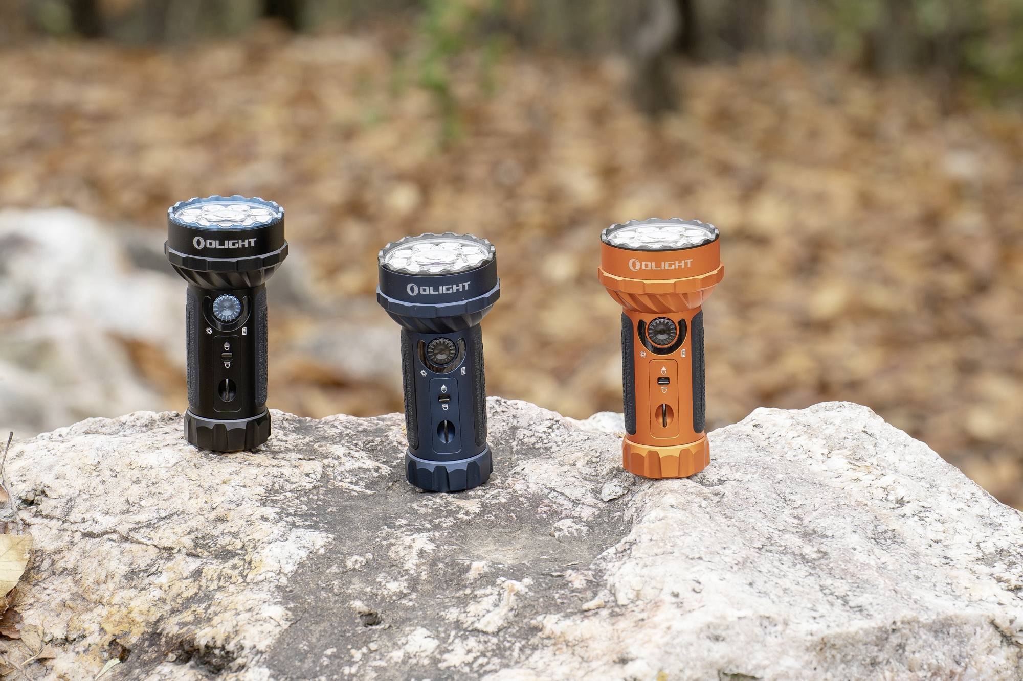 Drei Taschenlampen in verschiedenen Farben (schwarz, blau und orange) stehen auf einem großen Felsen, Hintergrund sind unscharfe Blätter.