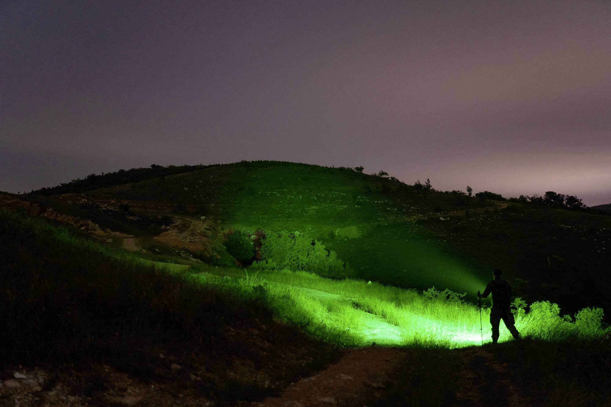Person mit grünem Spotlight beleuchtet nächtliche Hügellandschaft. Dunkler Himmel und diffuse Lichter am Horizont im Hintergrund.