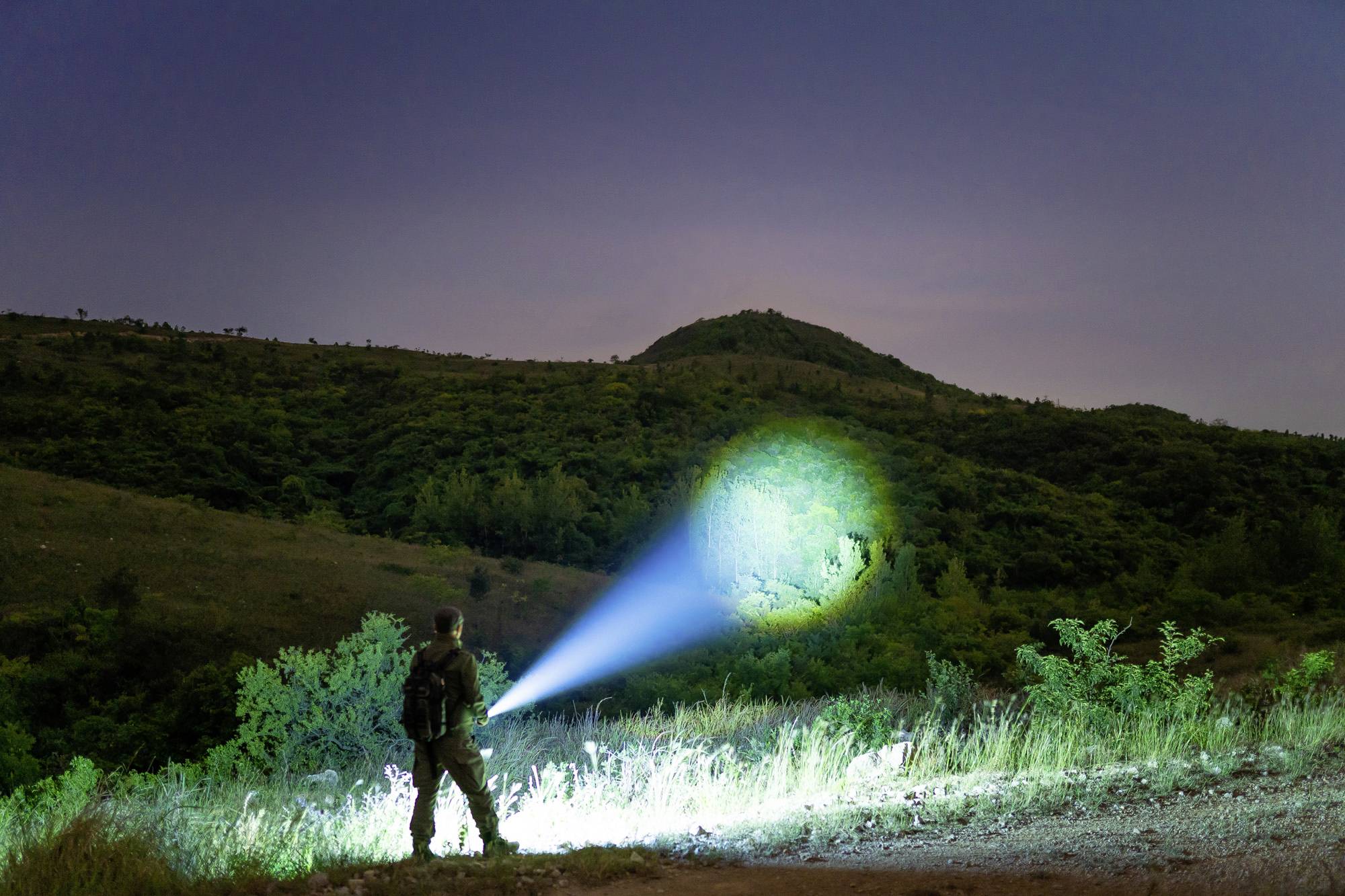 Eine Person steht nachts mit einer Taschenlampe in der Hand auf einem Feld und beleuchtet die angrenzende Hügellandschaft.