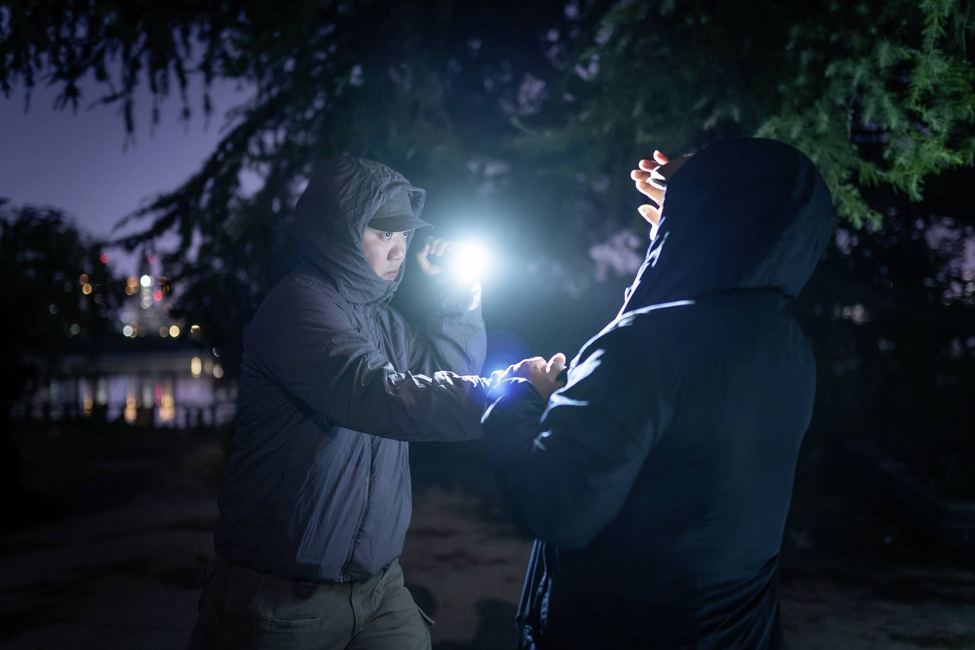 Zwei Personen in Jacken streiten sich im Dunkeln, eine Person hält eine Taschenlampe, die den anderen blendet.