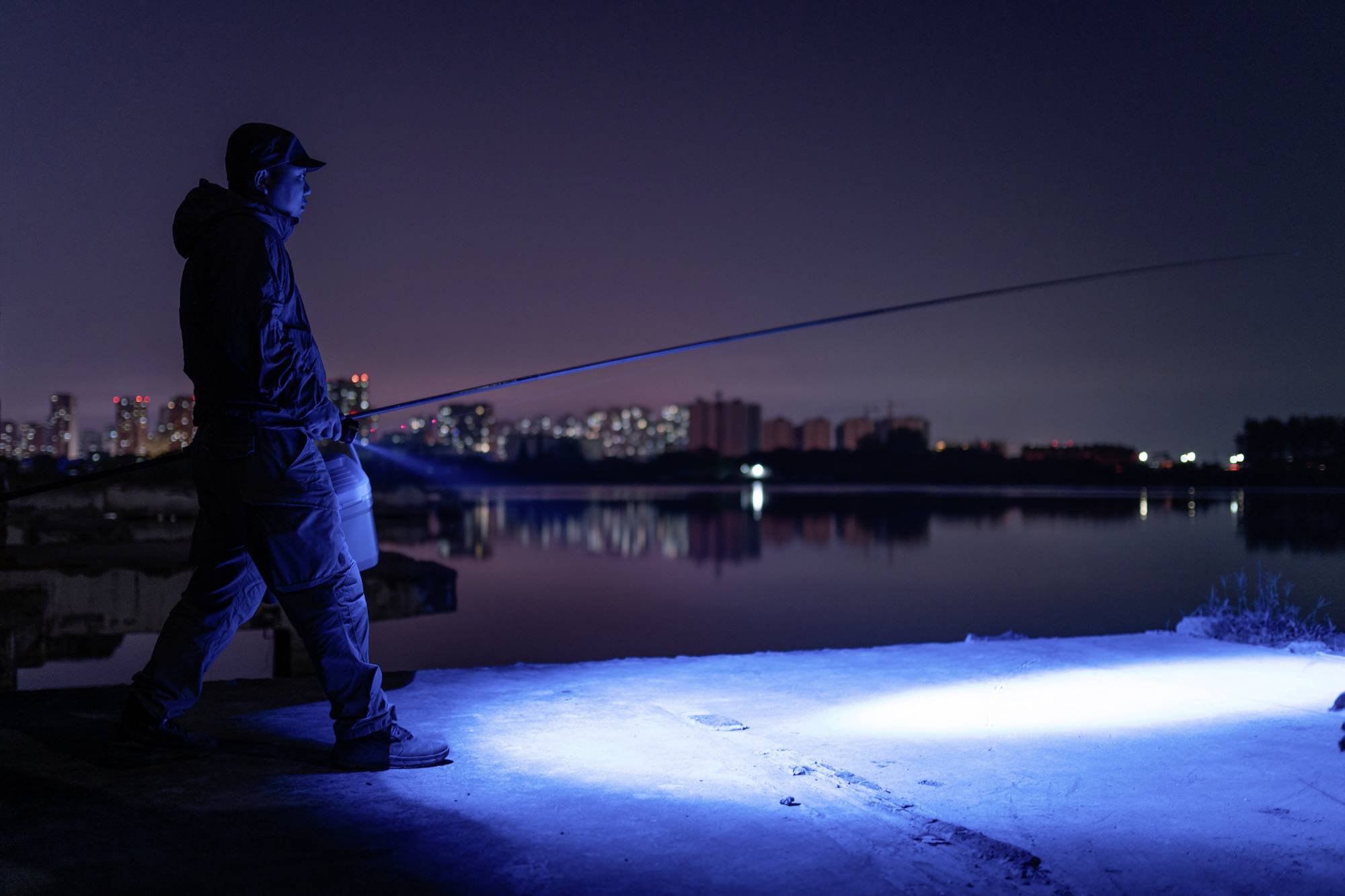 Eine Person angelt nachts an einem ruhigen Gewässer. Die Stadtlichter erhellen den Hintergrund, während eine Taschenlampe den Boden beleuchtet.