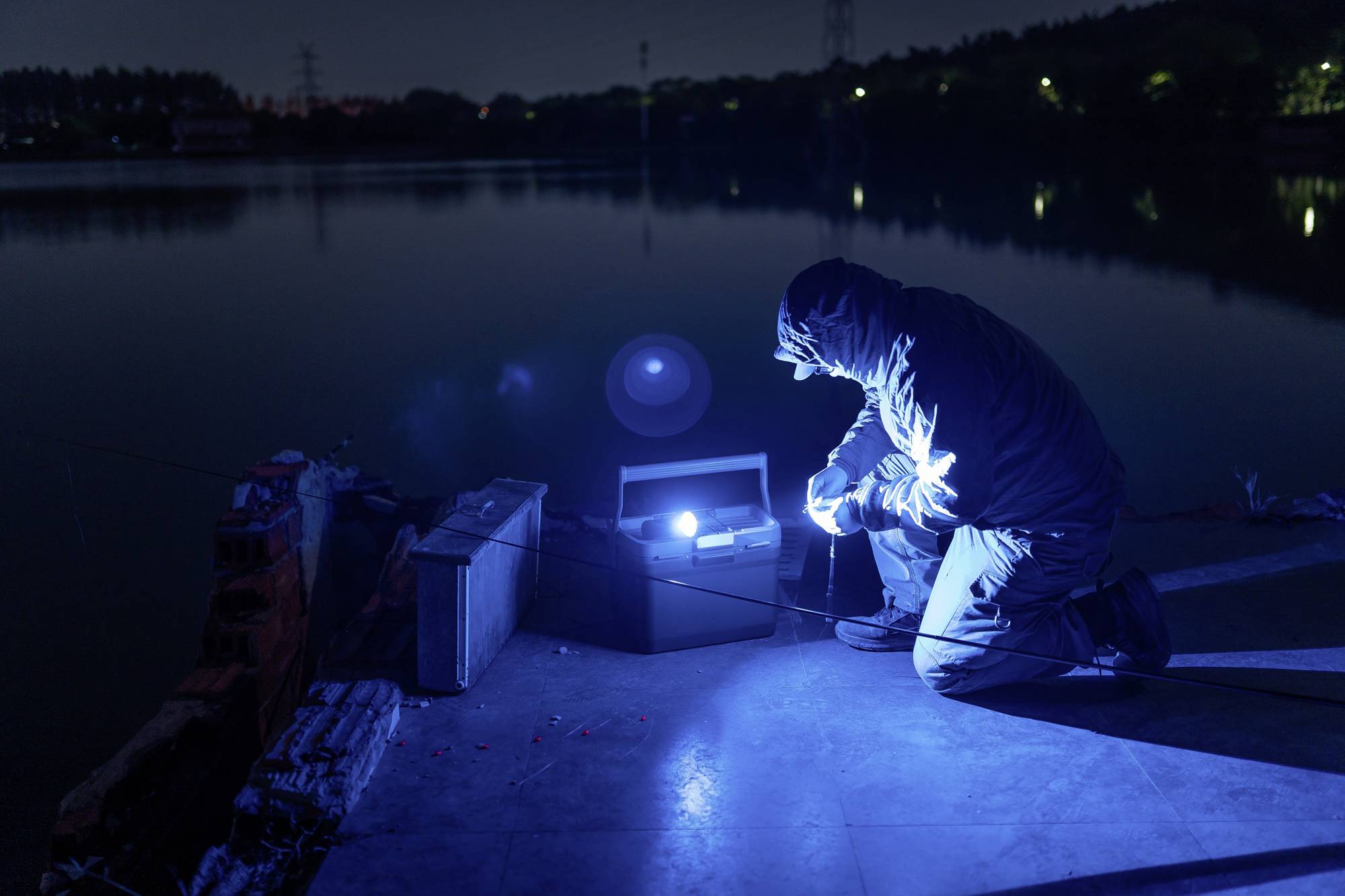 Eine Person kniet nachts neben einem Fluss und arbeitet mit einem blauen Licht auf technischem Gerät. Hintergrund zeigt dunkle Bäume.