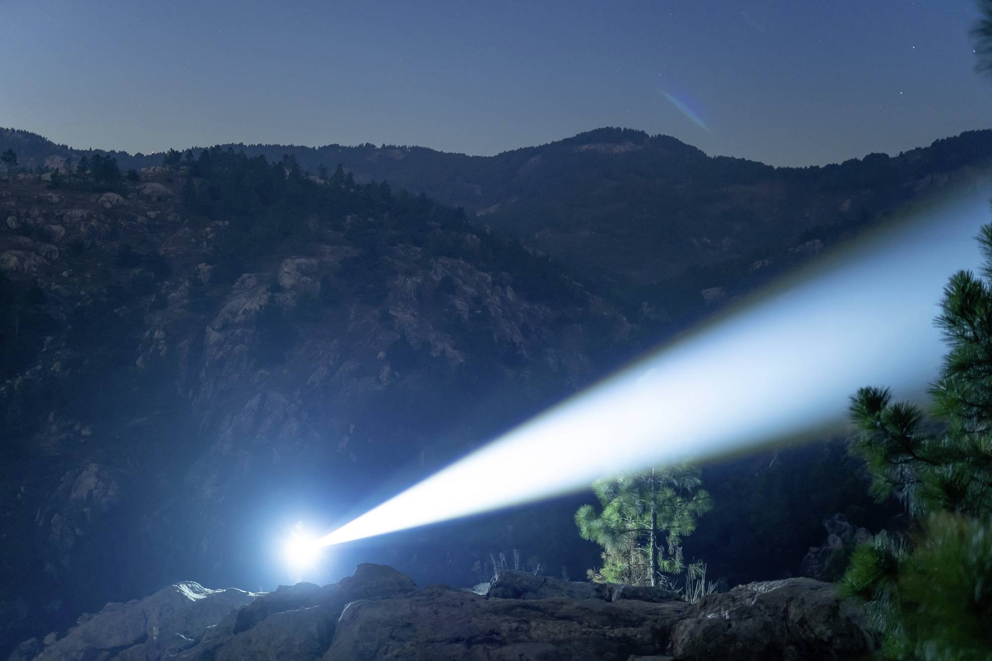 Ein heller Lichtstrahl eines Scheinwerfers durchleuchtet die Nacht in einer bergigen Landschaft mit Bäumen im Vordergrund.
