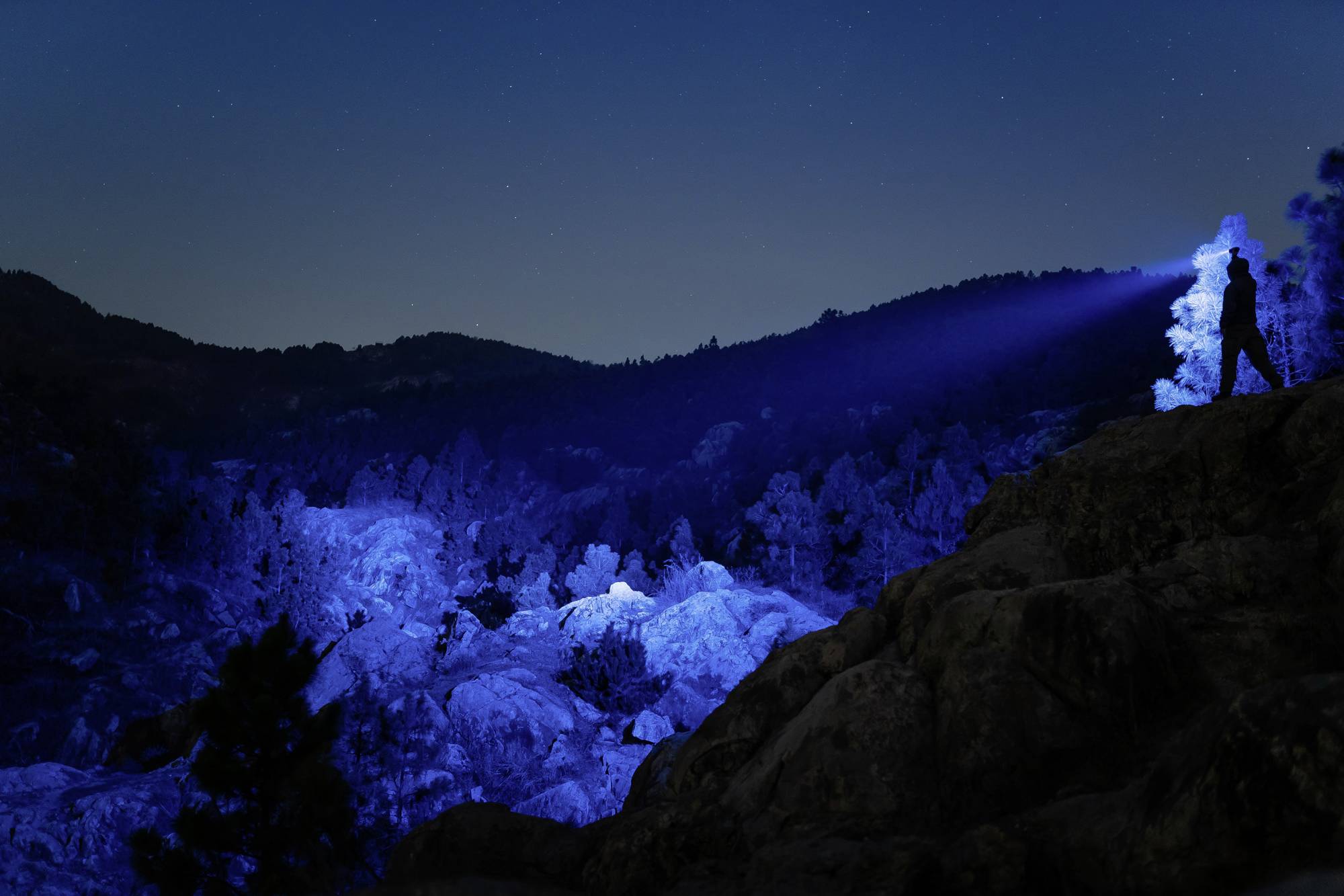 Person mit Taschenlampe auf einem Felsen beleuchtet nächtliche Landschaft, Berge im Hintergrund.
